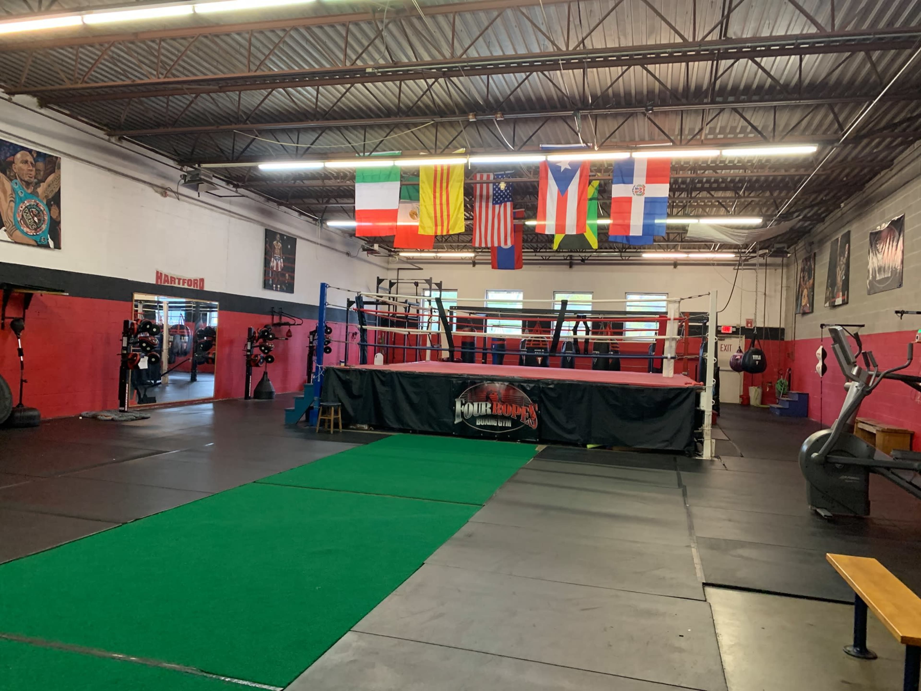 A boxing gym featuring a central ring, exercise equipment, and various flags hanging from the ceiling.