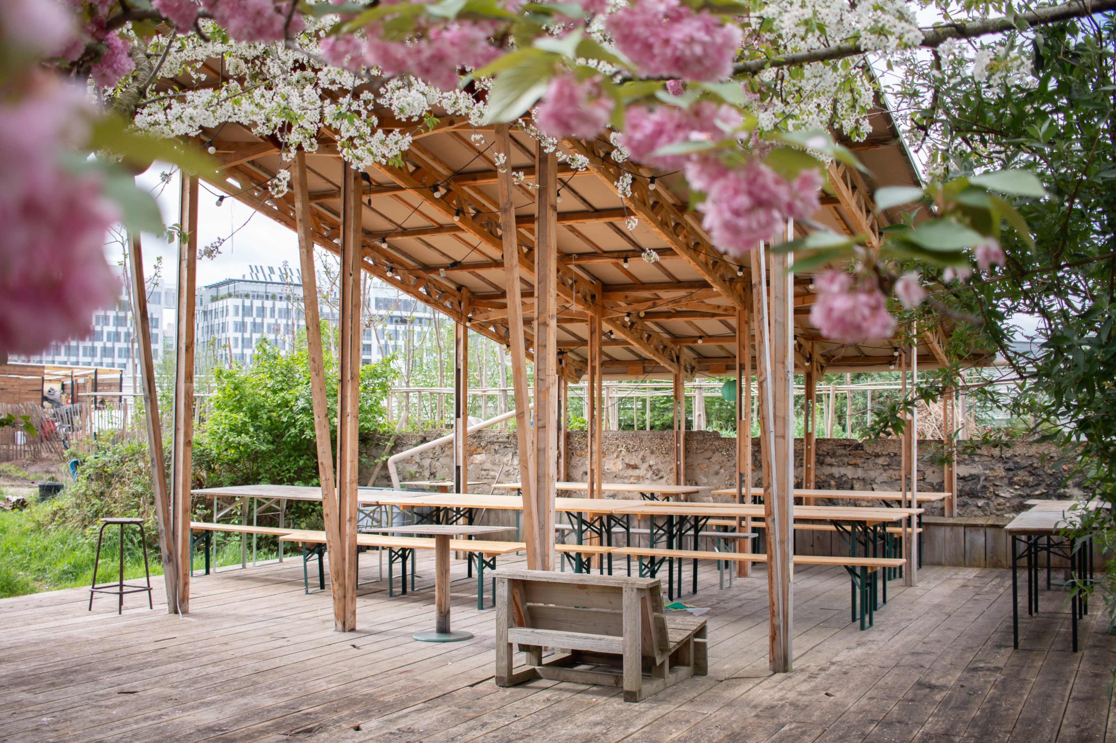 A wooden pavilion with tables and chairs is surrounded by greenery and flowering trees.