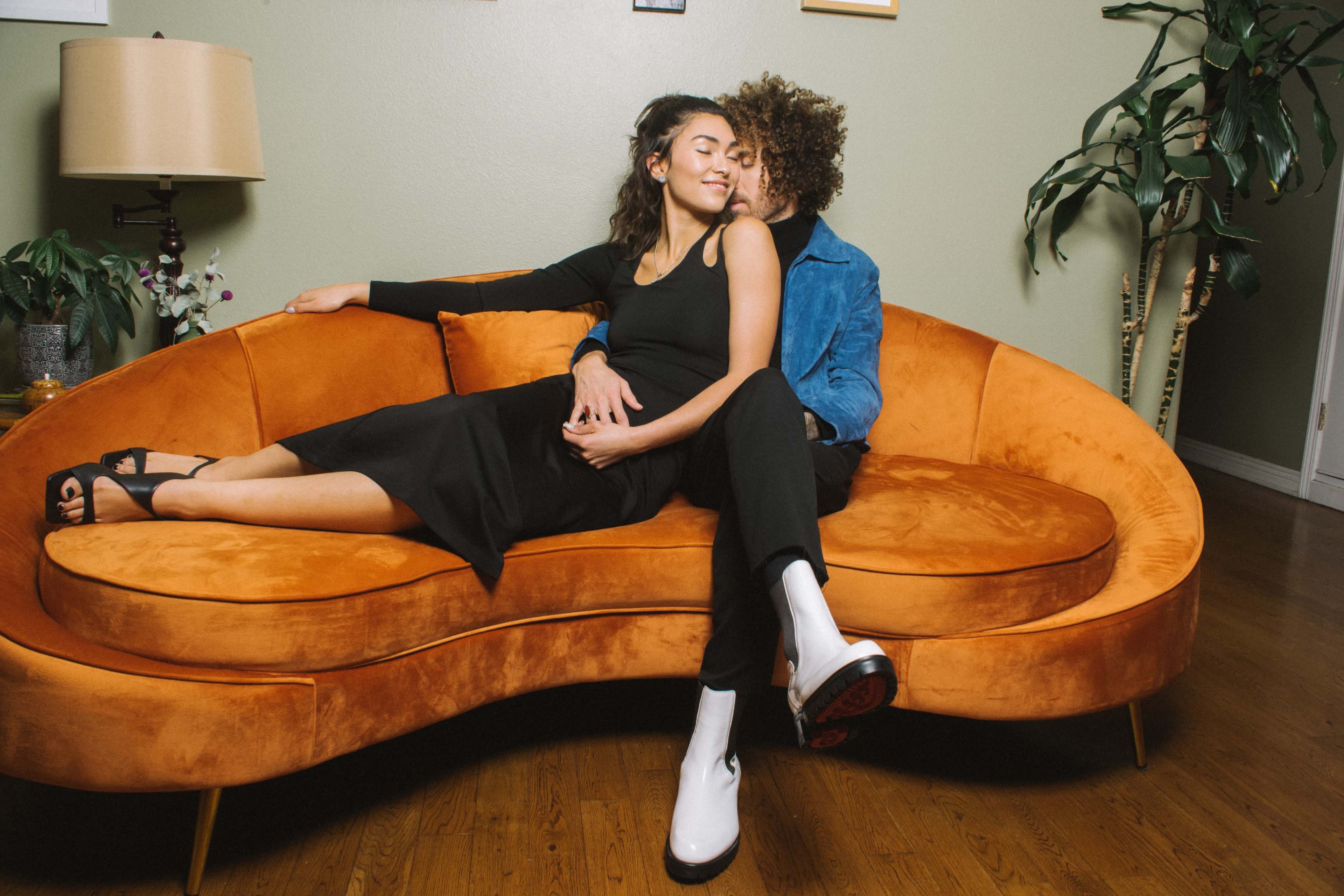 A woman in a black dress lounges on an orange sofa while a man in a blue jacket sits close behind her.