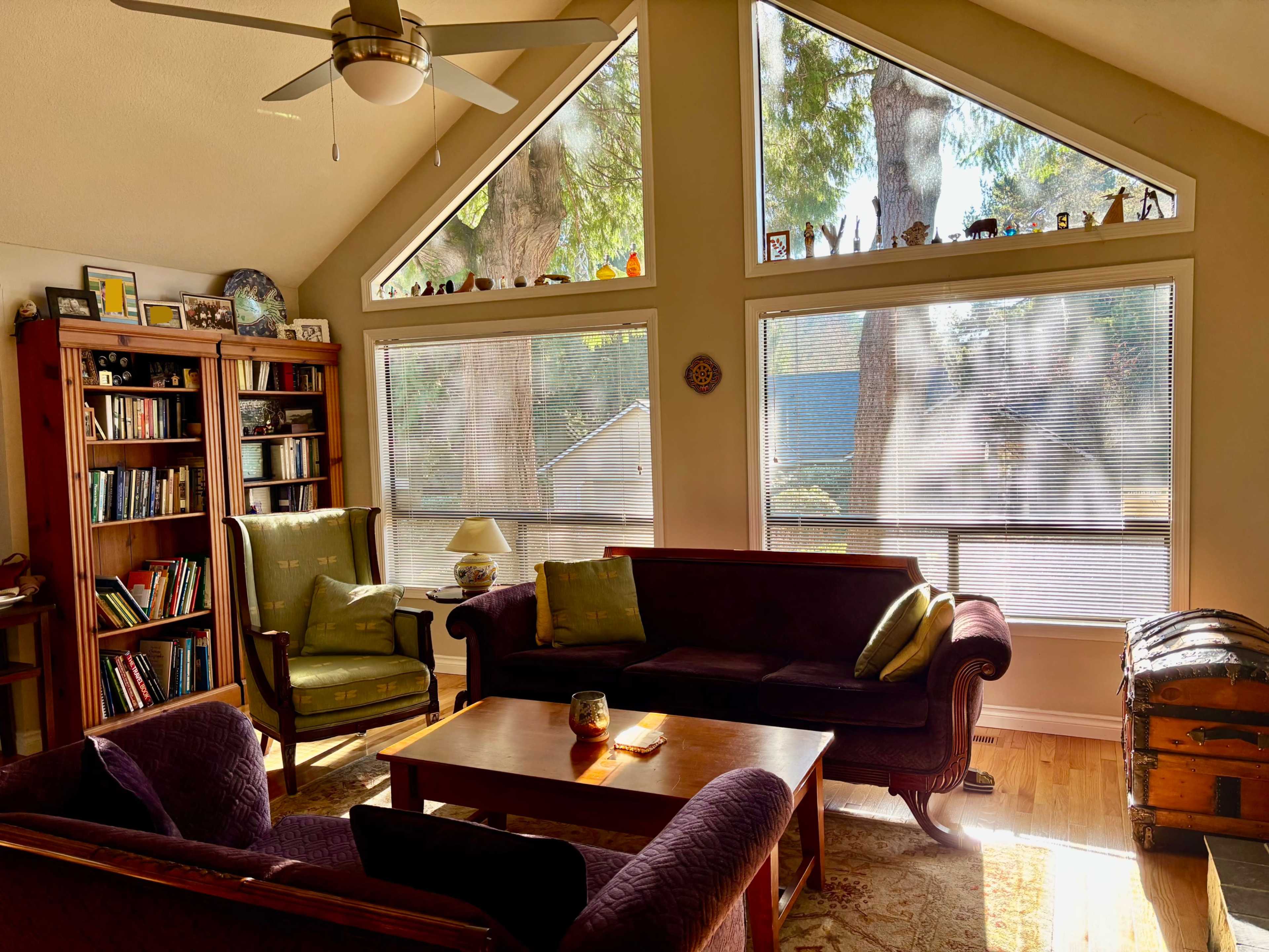 A cozy living room features a purple sofa and chairs, a wooden coffee table, a bookshelf, and large windows that allow sunlight to illuminate the space.