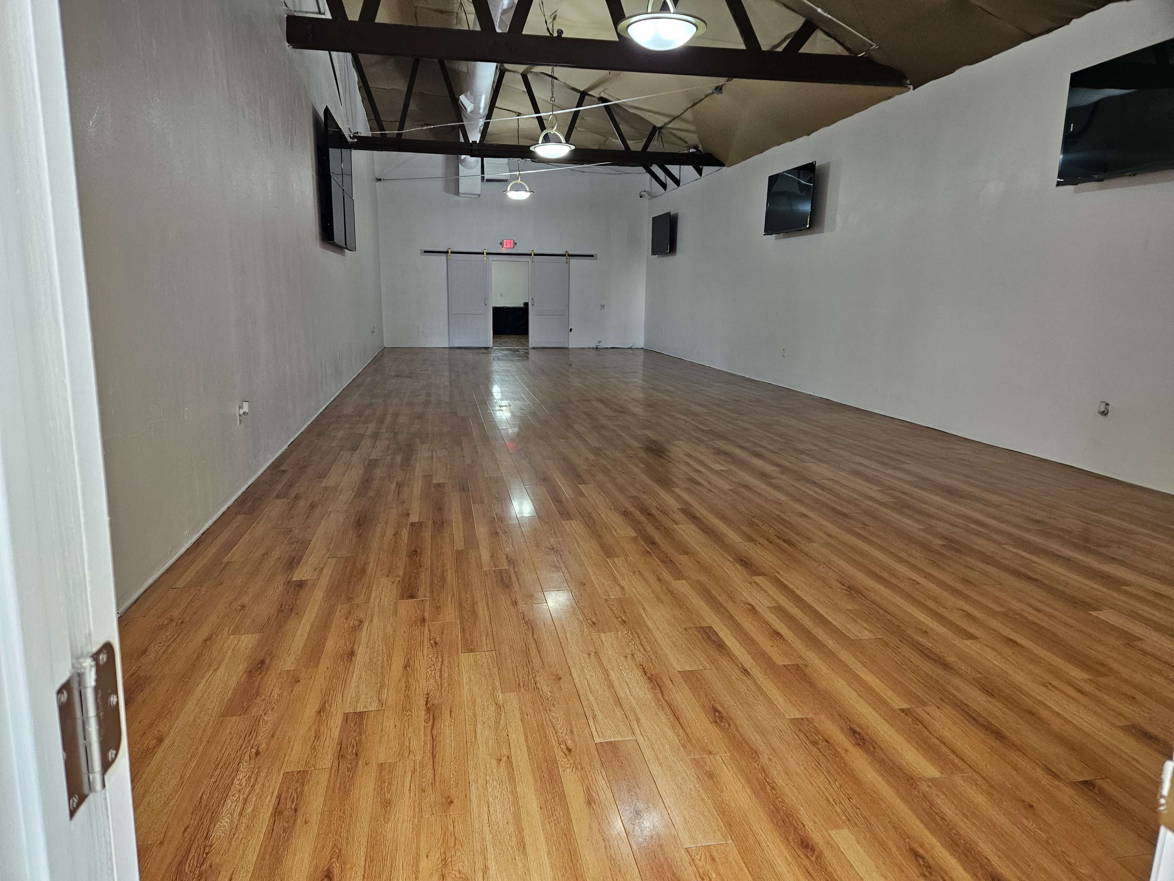 A spacious, empty room with wooden flooring, high ceilings, and multiple wall-mounted screens.