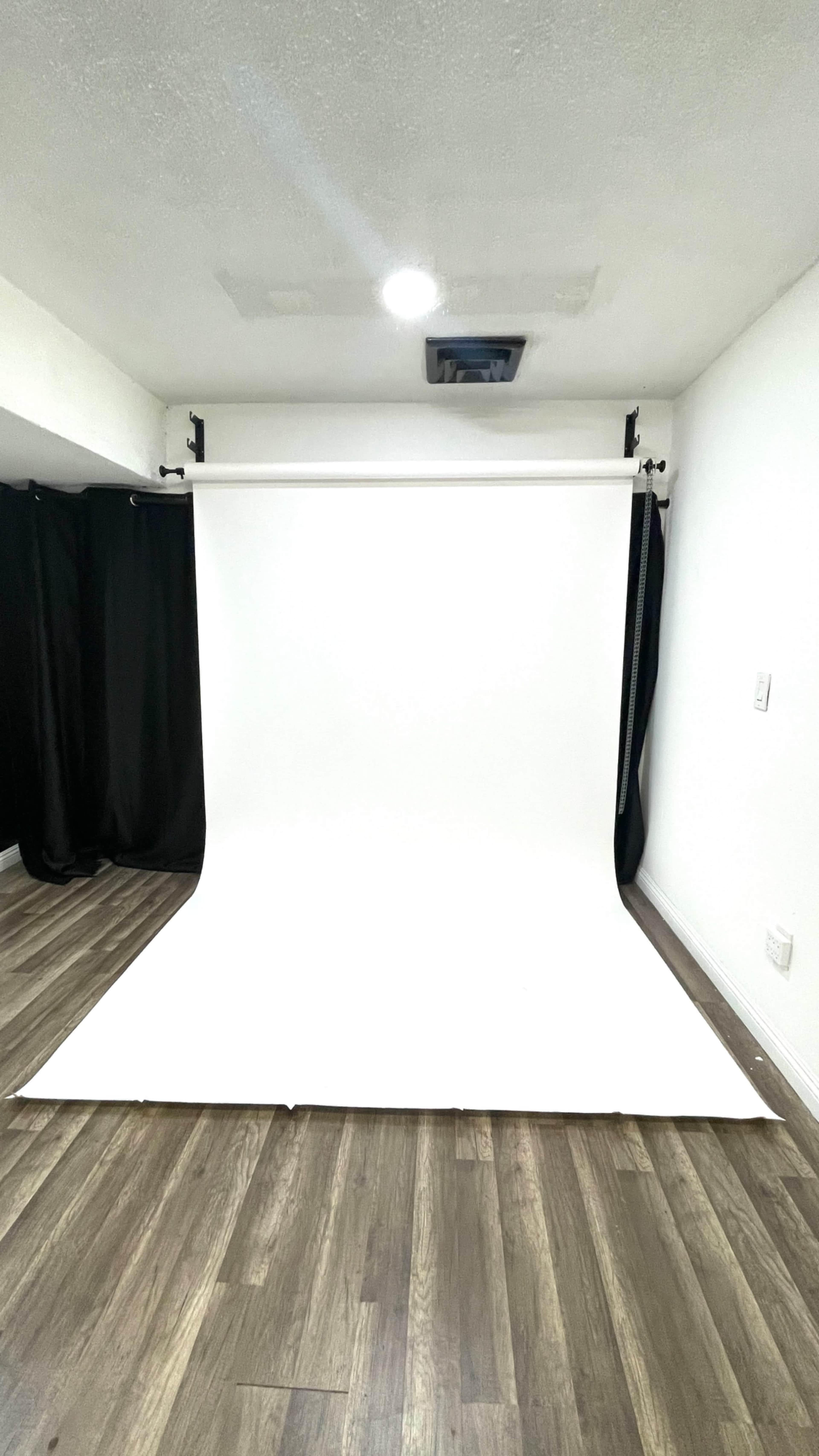 The image shows an empty photo studio with a white backdrop set against a wooden floor and black curtains on one side.