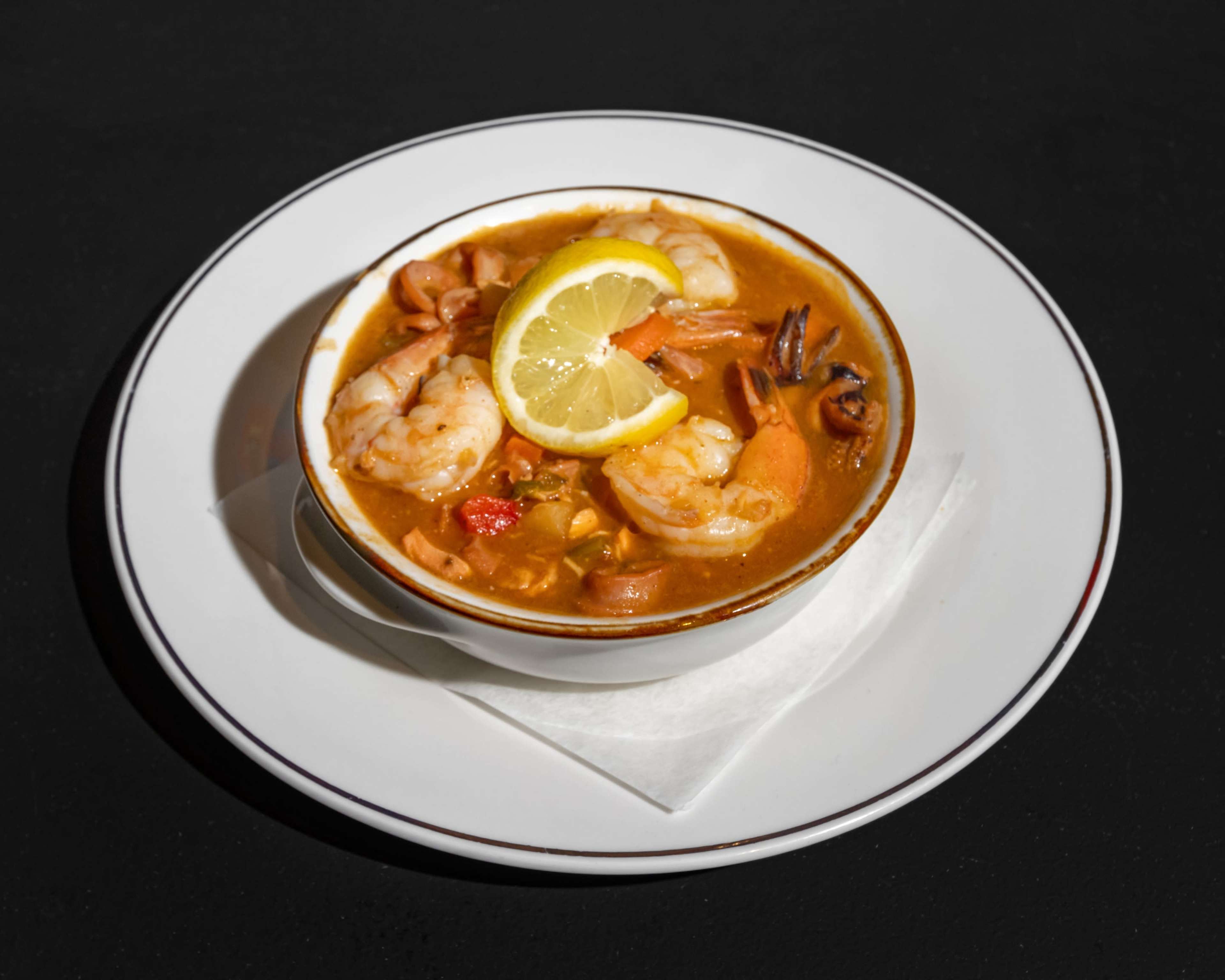 A bowl of seafood soup with shrimp, vegetables, and a slice of lemon sits on a white plate.