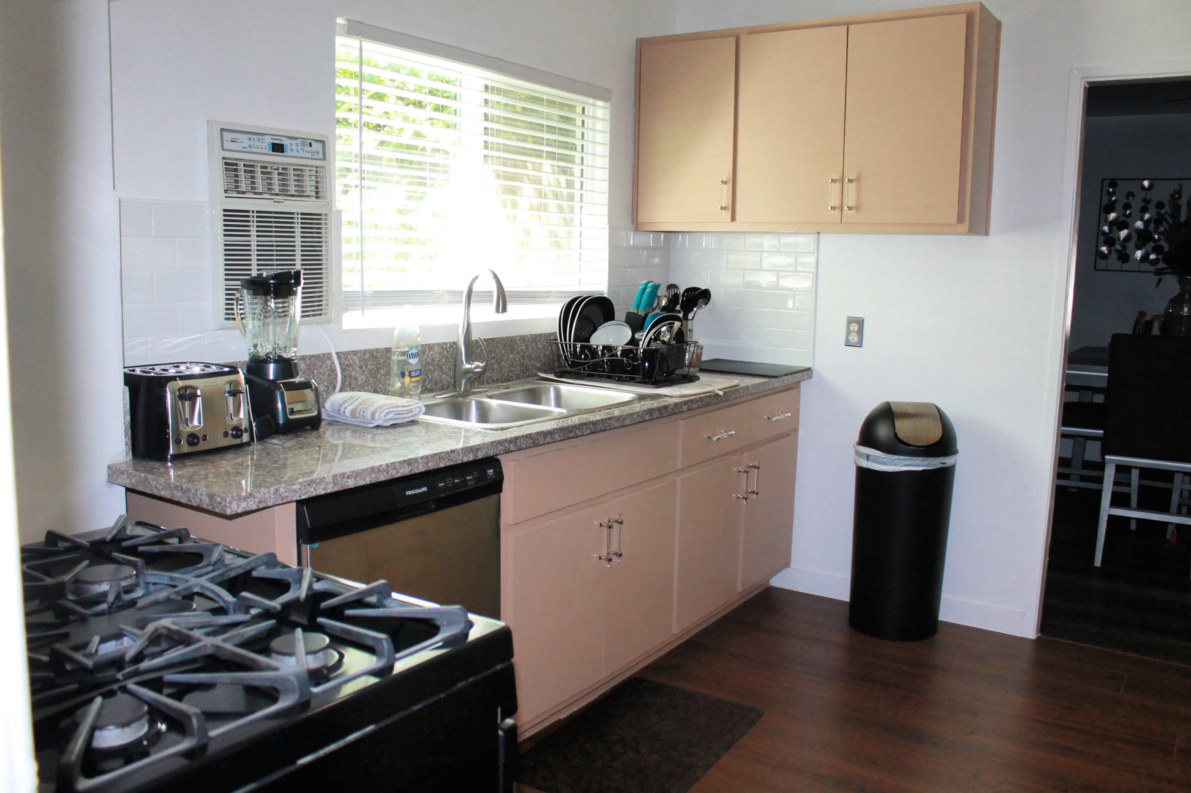 The image shows a modern kitchen with beige cabinets, a double sink, a stove, a dishwasher, and a trash can, situated next to a window.