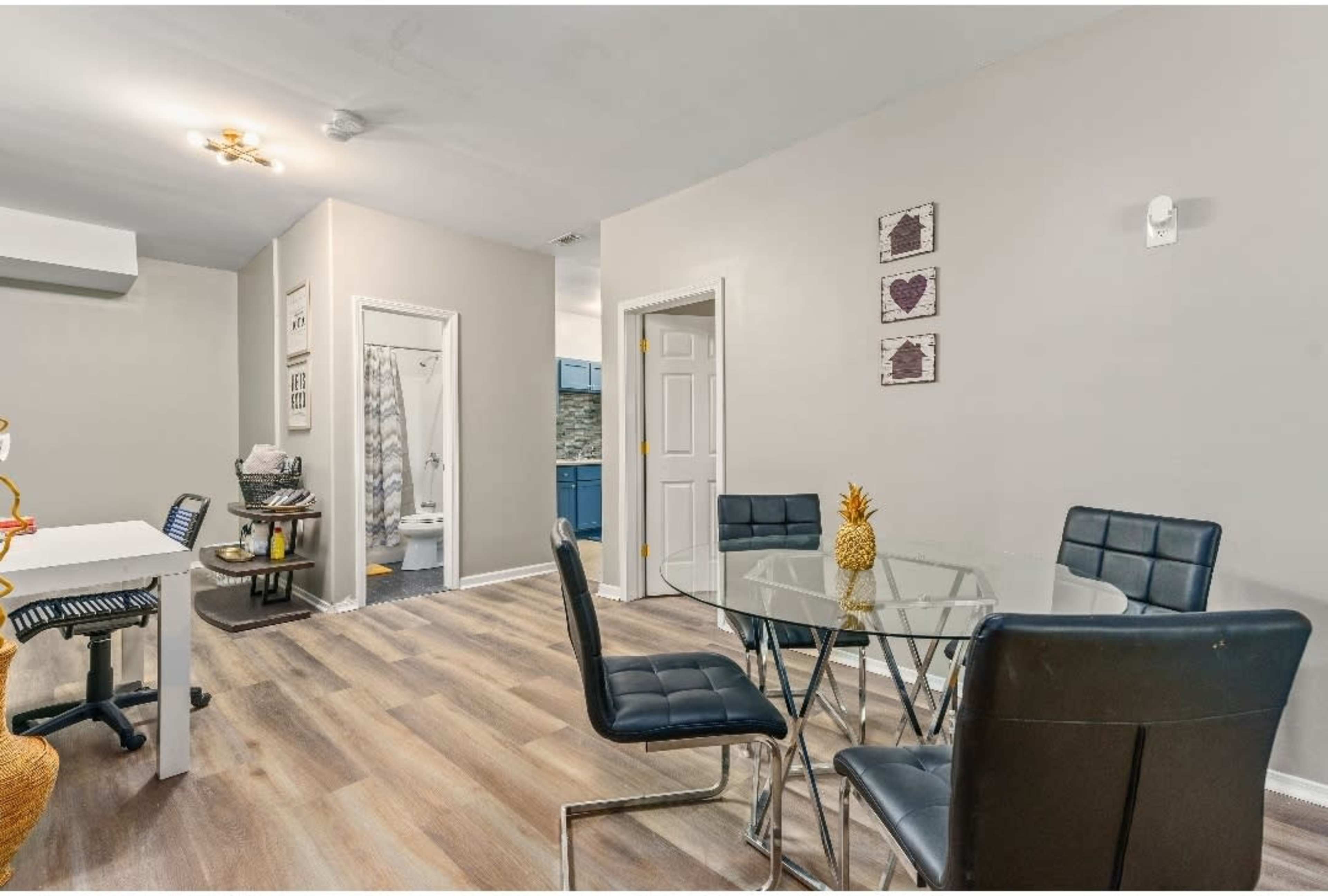 A dining area features a glass table surrounded by black chairs, with a view of a bathroom and kitchen in the background.