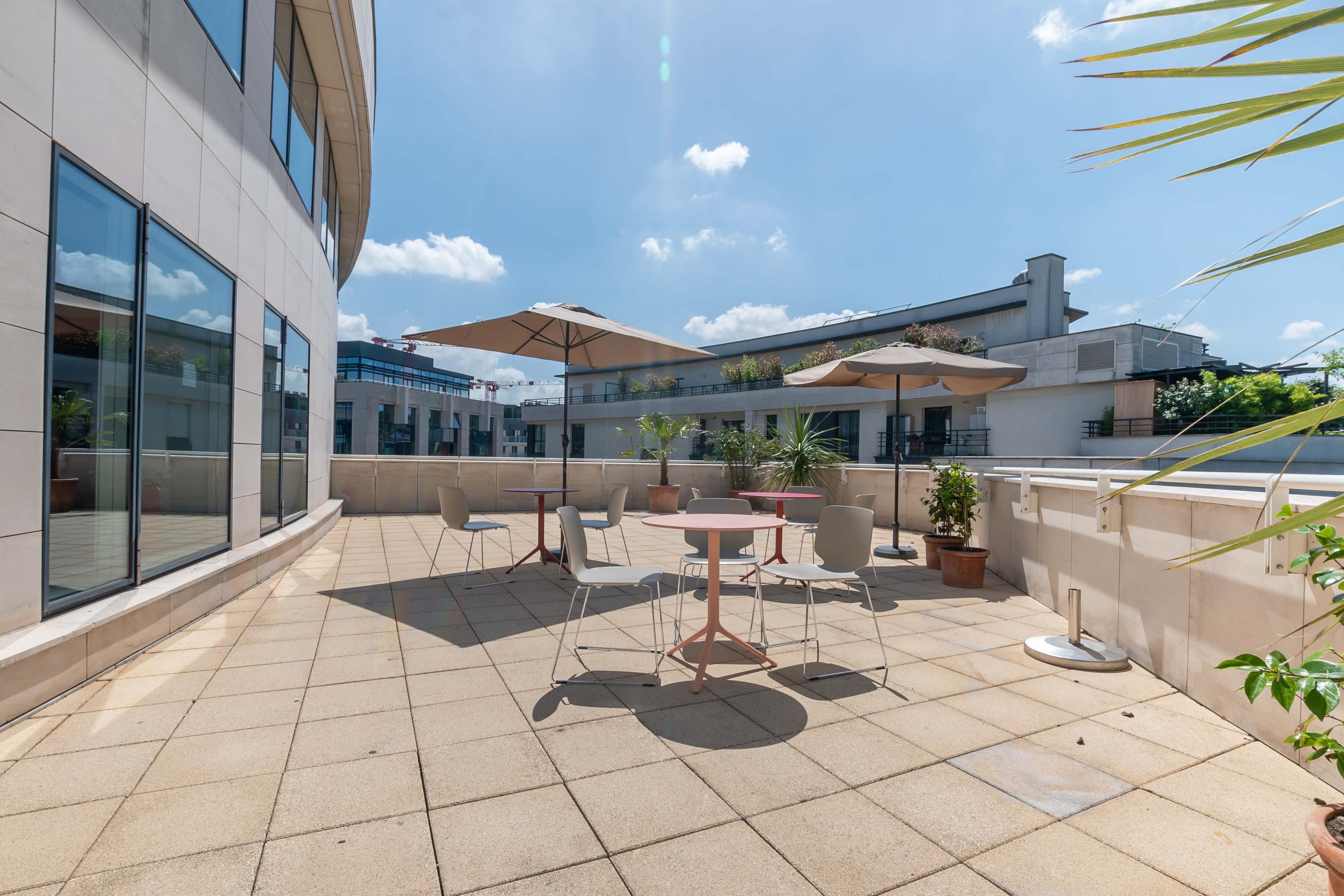 A spacious outdoor terrace with several tables and umbrellas, surrounded by windows of a modern building.