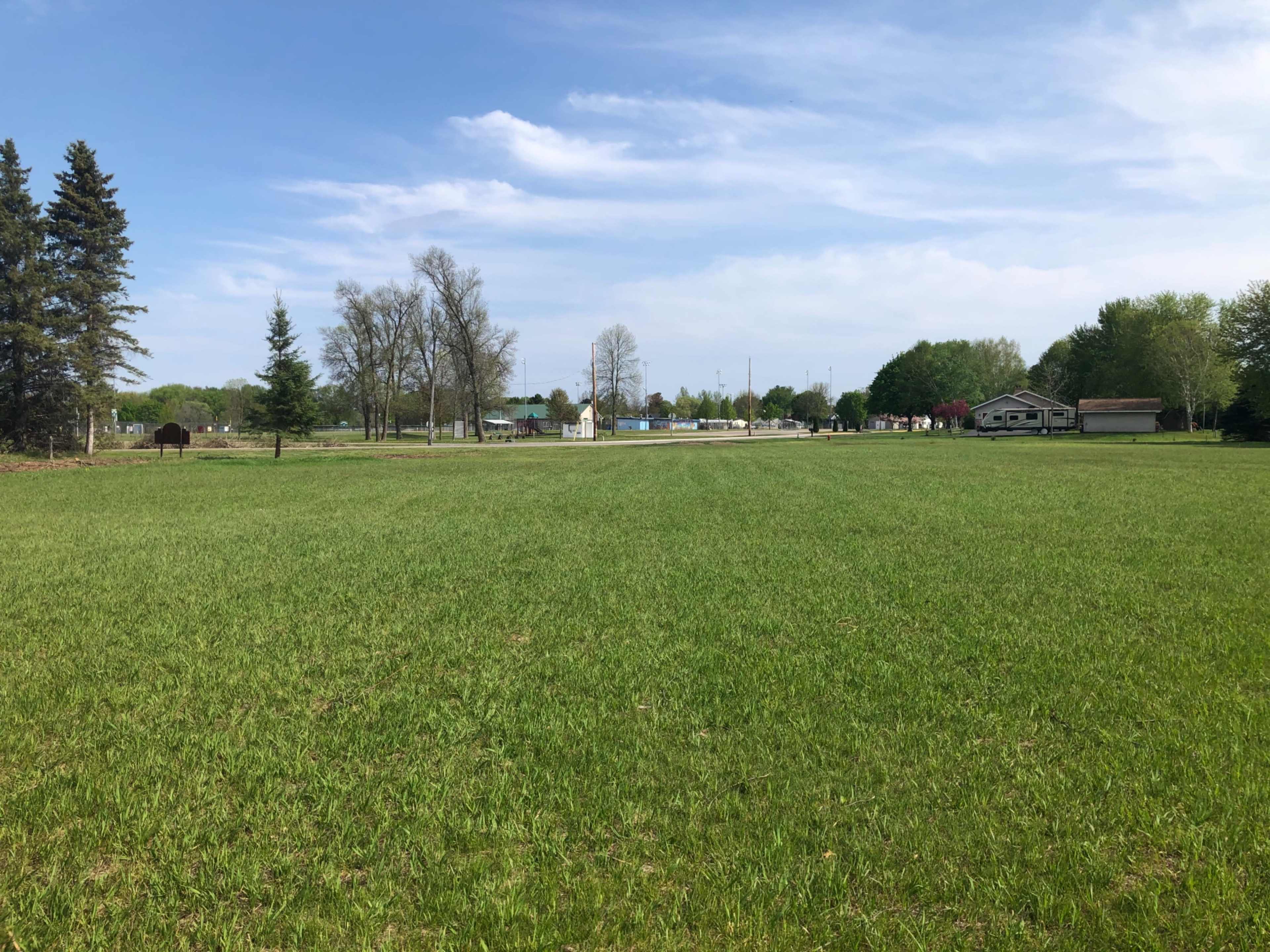 A wide, grassy field stretches toward a distant row of trees and buildings under a partly cloudy sky.