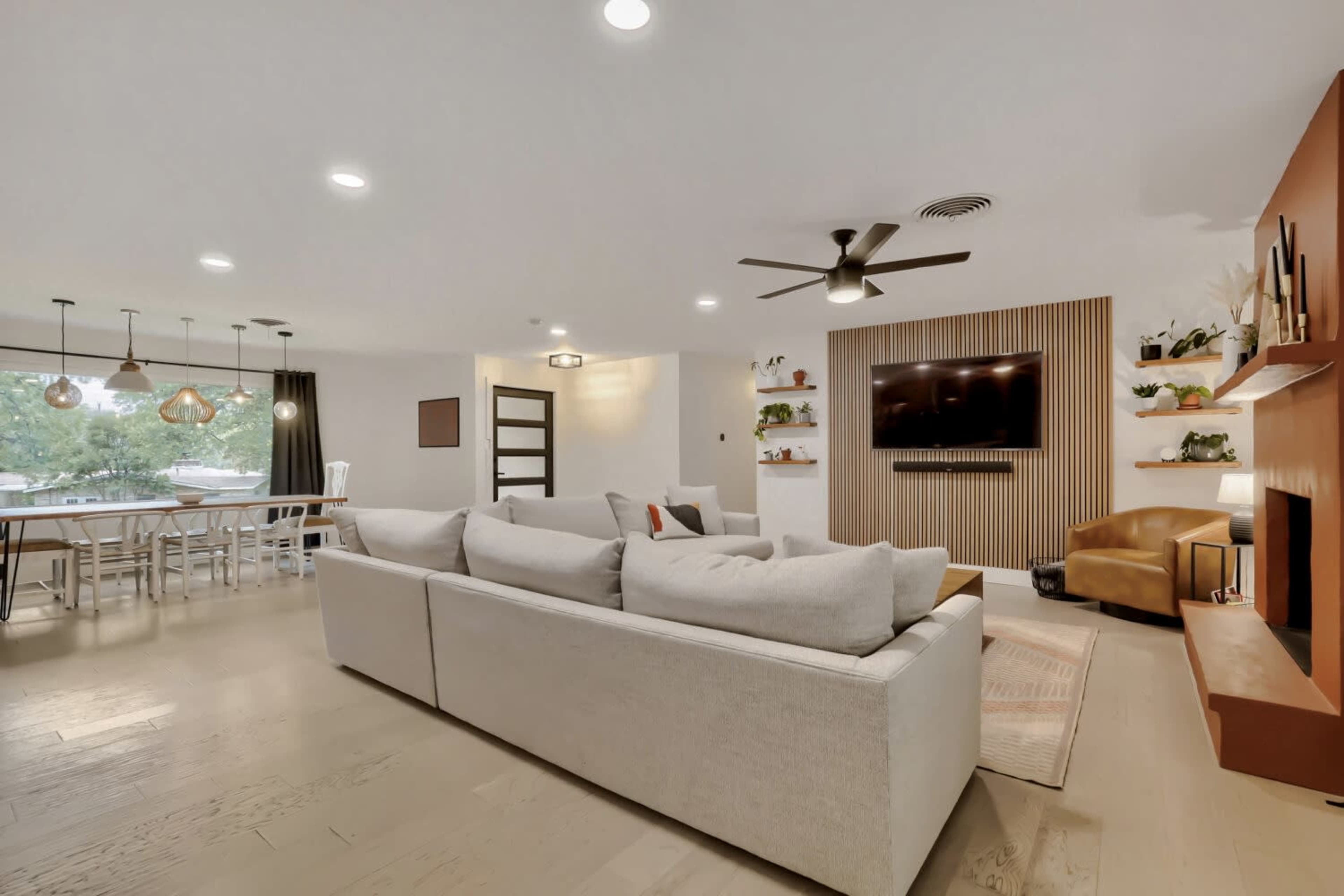 The image features a modern living room with a light-colored sofa, a wall-mounted television, and a dining area with a large table and pendant lighting.