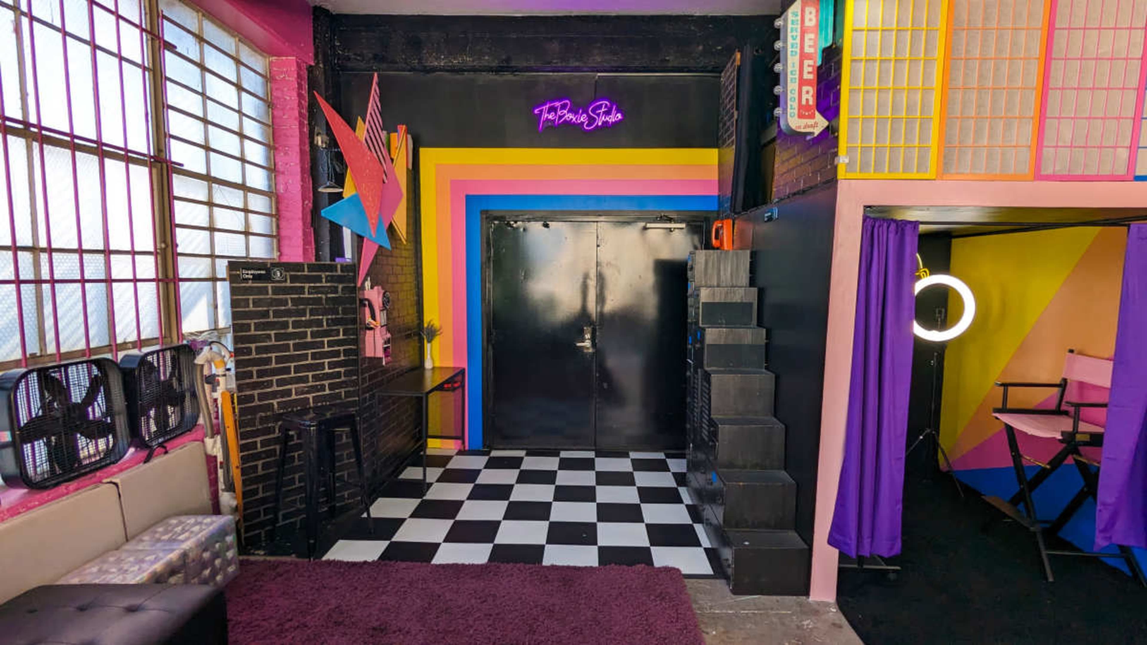 A vibrant studio space featuring colorful walls, a checkered floor, and a black door at the back, with various decorations and furniture surrounding the area.