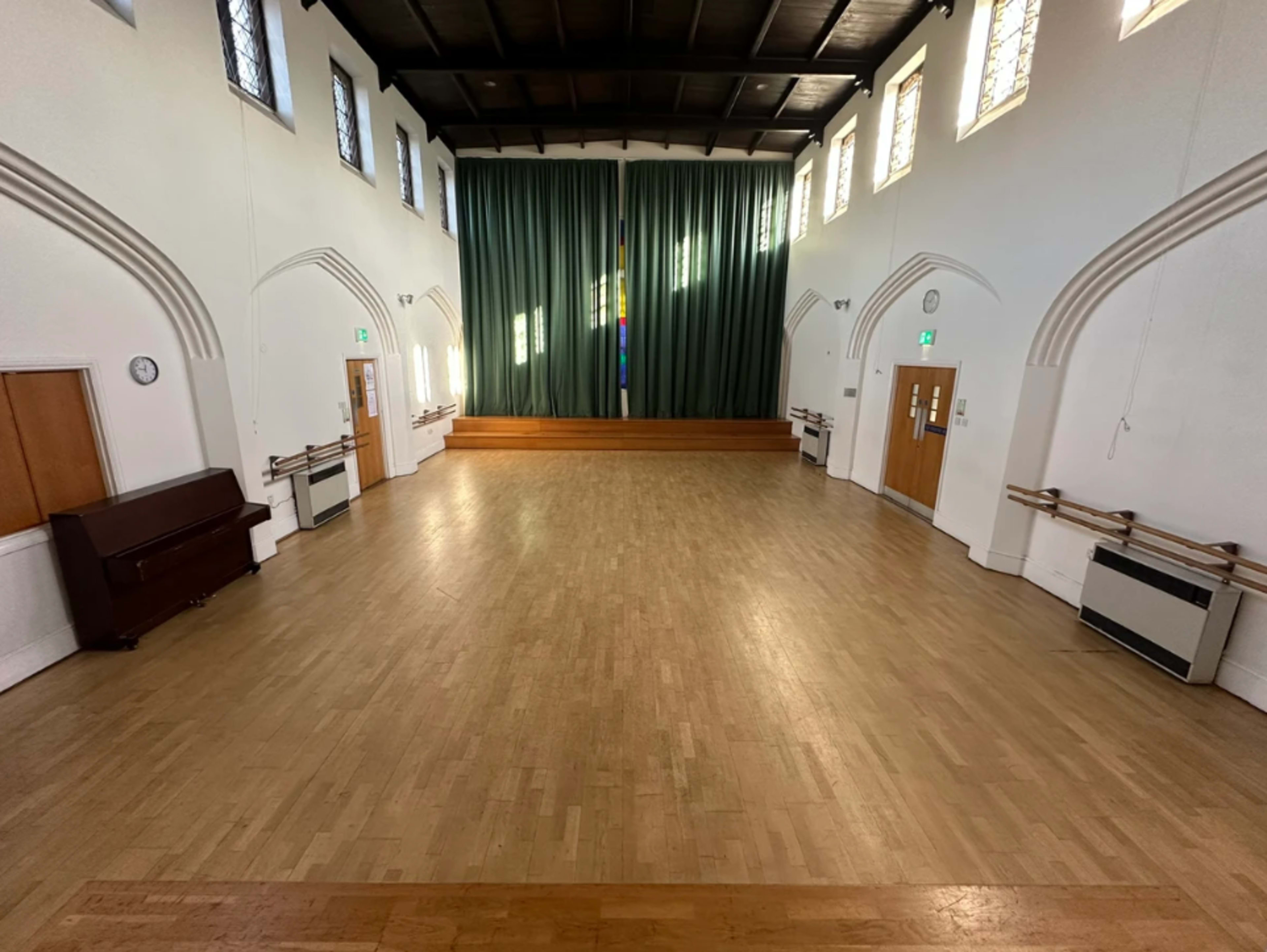 The image shows a spacious hall with a wooden floor, large windows, and green curtains at the front, along with a piano against one wall.