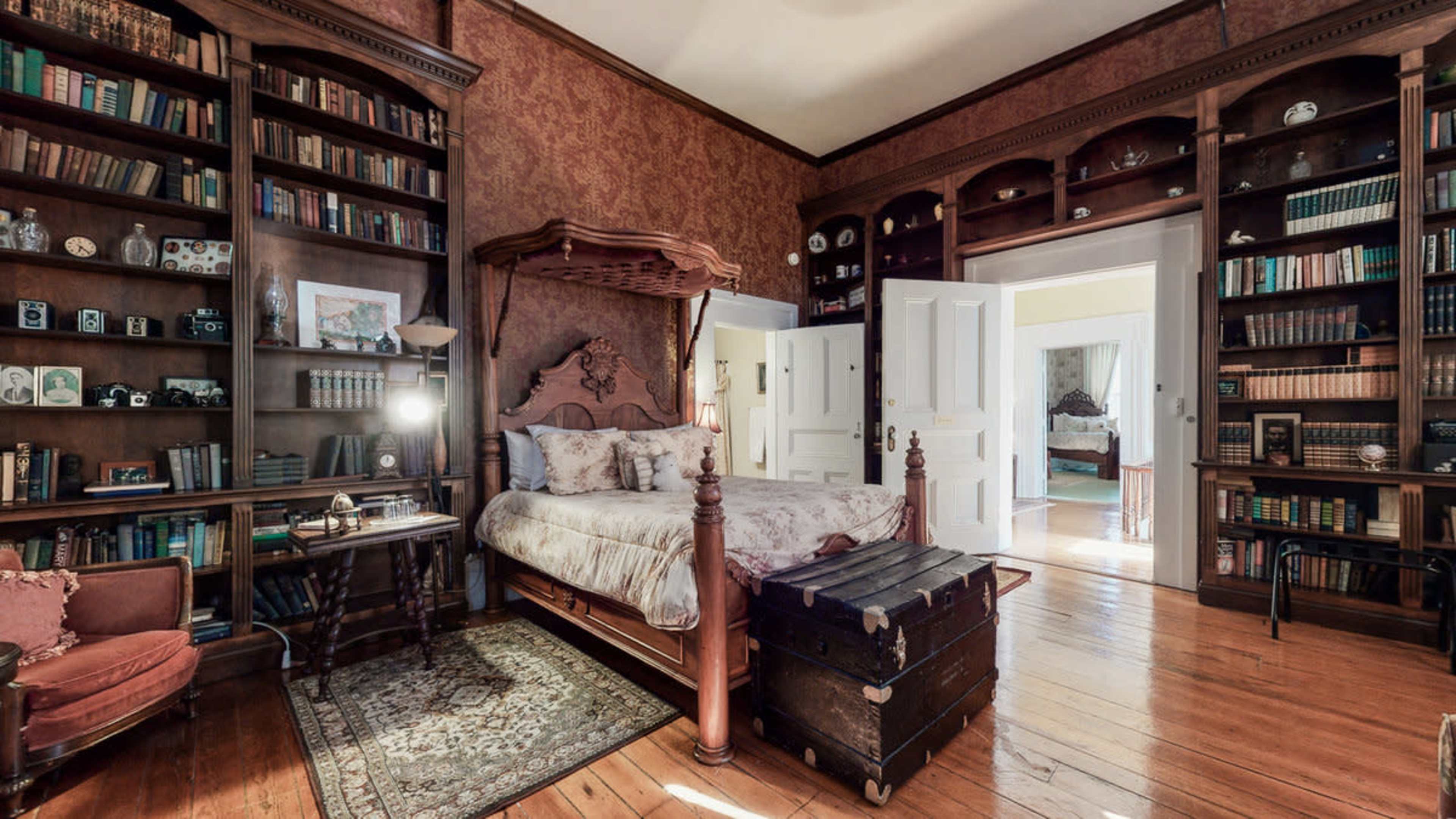 A bedroom with a four-poster bed, surrounded by tall bookshelves filled with books and decorative items, and a vintage trunk at the foot of the bed.