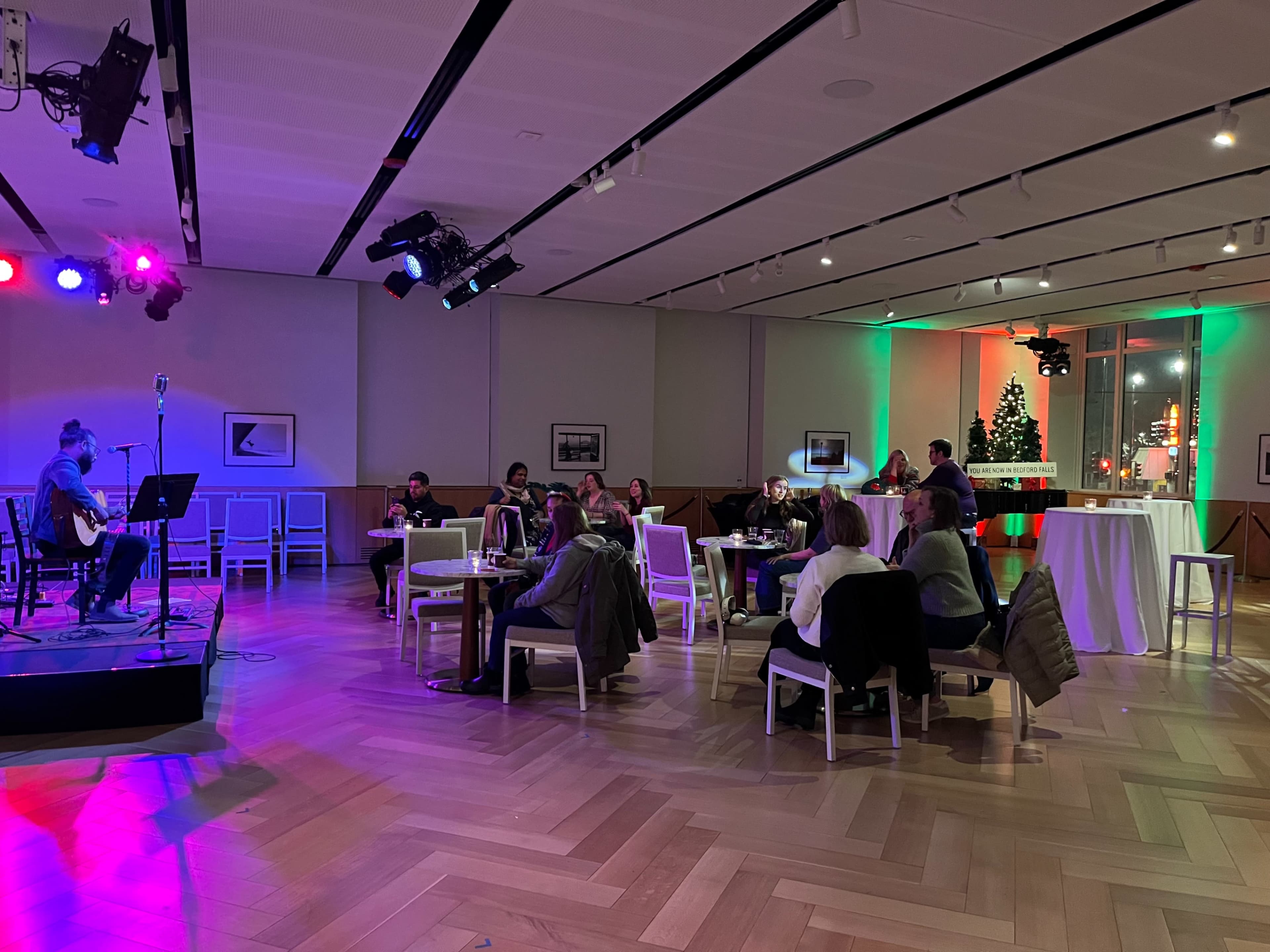 A musician performs on a stage while an audience seated at tables enjoys the evening in a well-lit venue adorned with colorful lights and decorations.