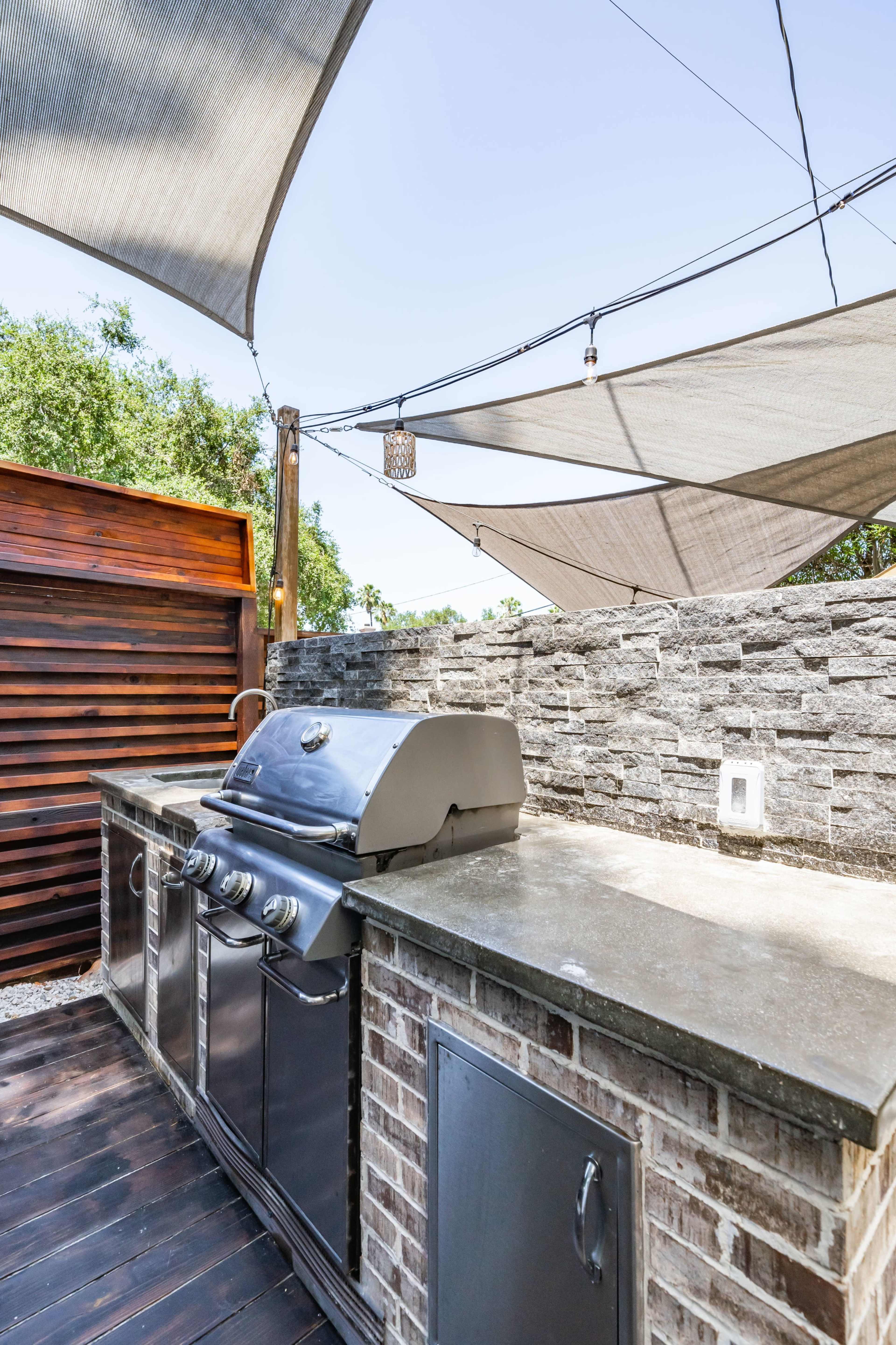 The image shows an outdoor kitchen area featuring a stainless steel grill and a stone countertop, surrounded by wooden privacy slats and shaded by fabric canopies.