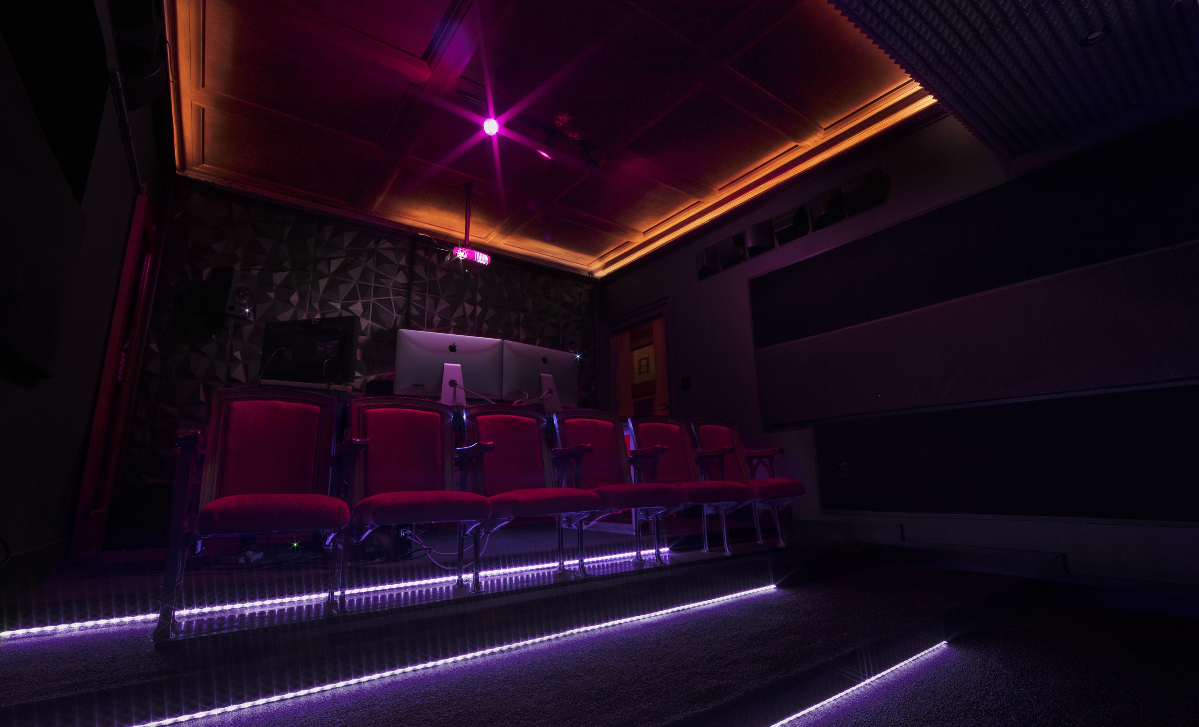 A dimly lit theater room features several red seats arranged in front of a control area, with purple lighting illuminating the space.