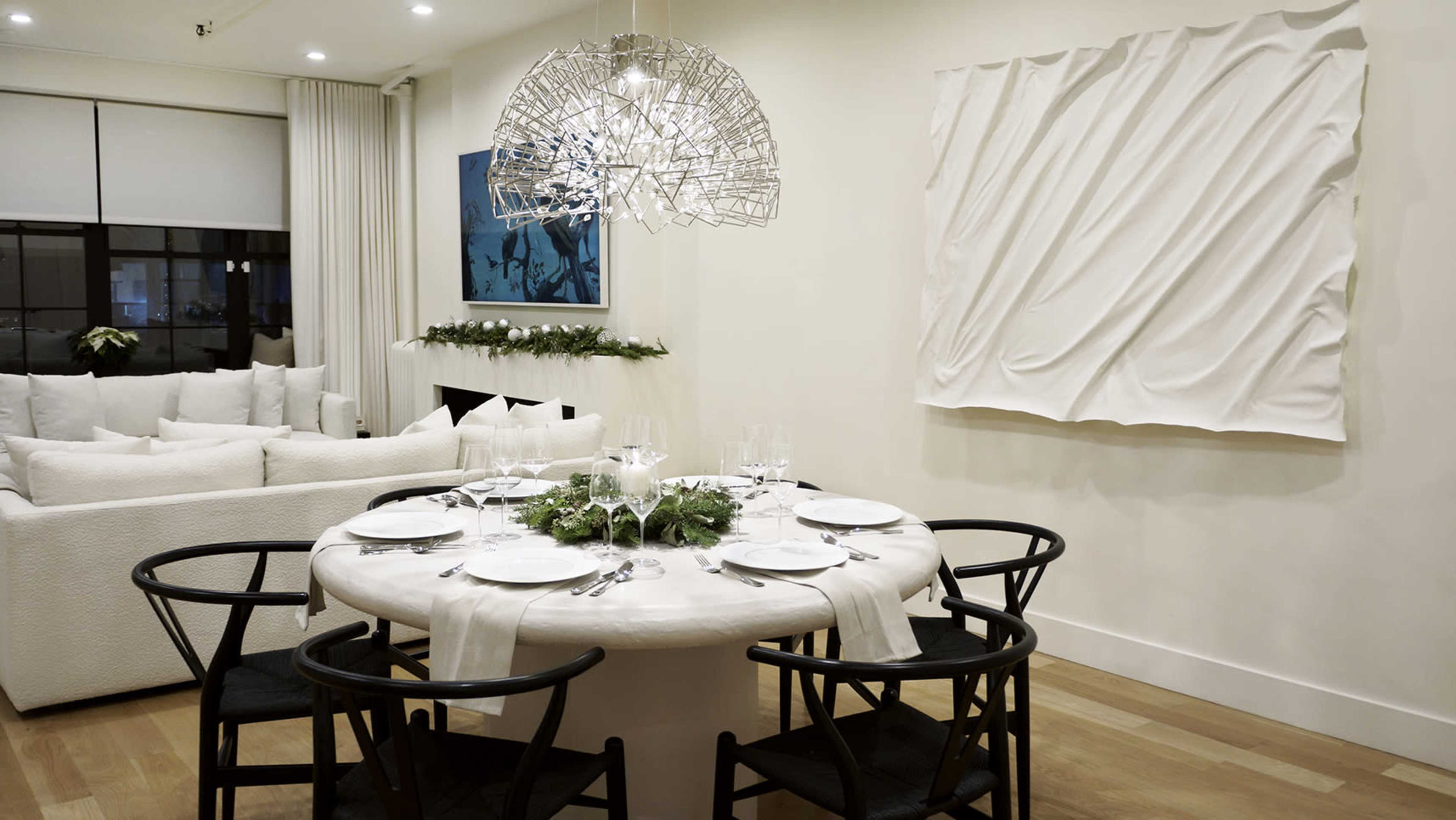 A round dining table is elegantly set for a meal, surrounded by black chairs, with a modern art piece and a plush sofa in the background.