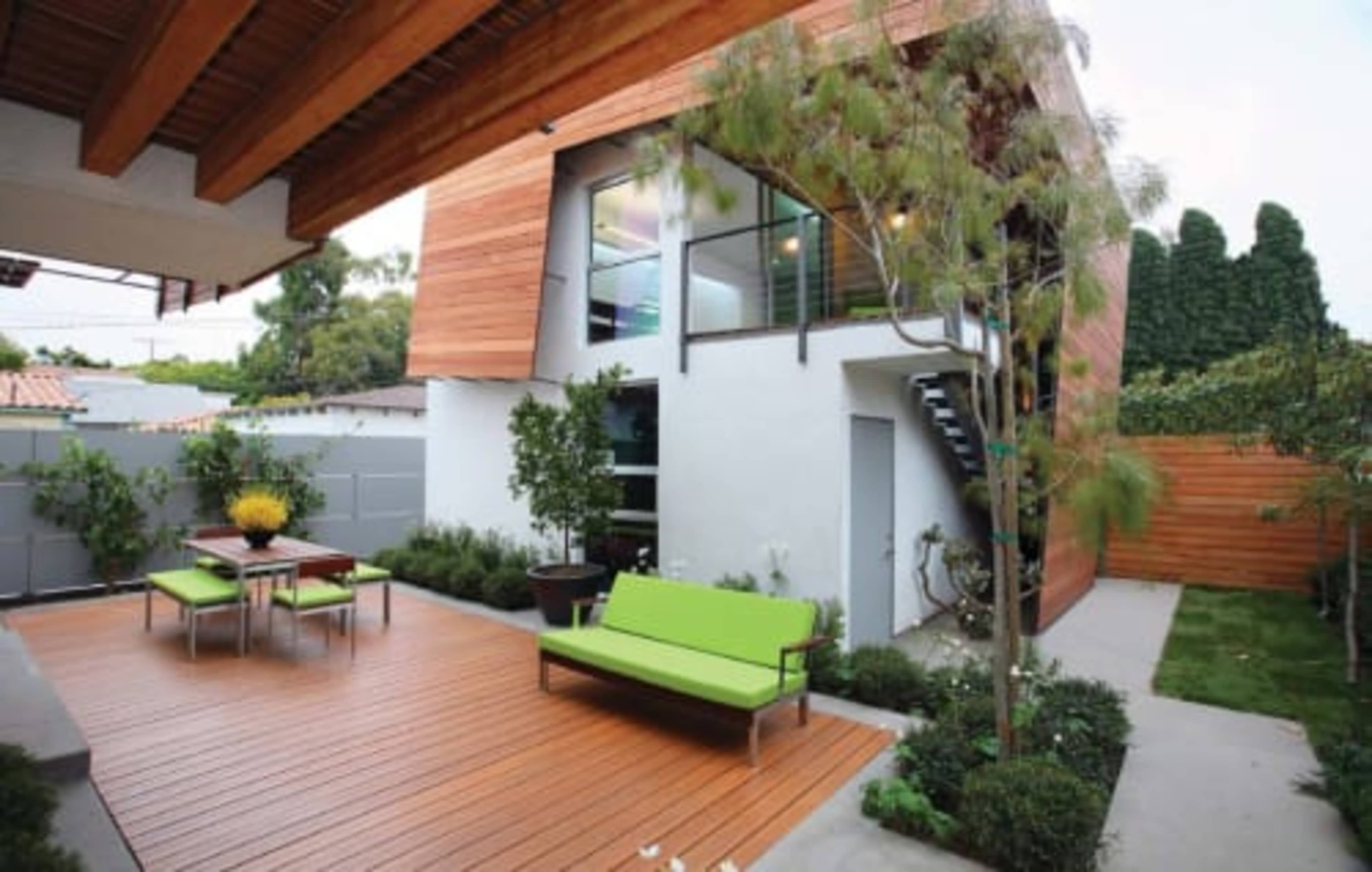 The image shows a modern two-story house with a wooden facade surrounded by a landscaped patio featuring wooden decking, green seating, and decorative plants.