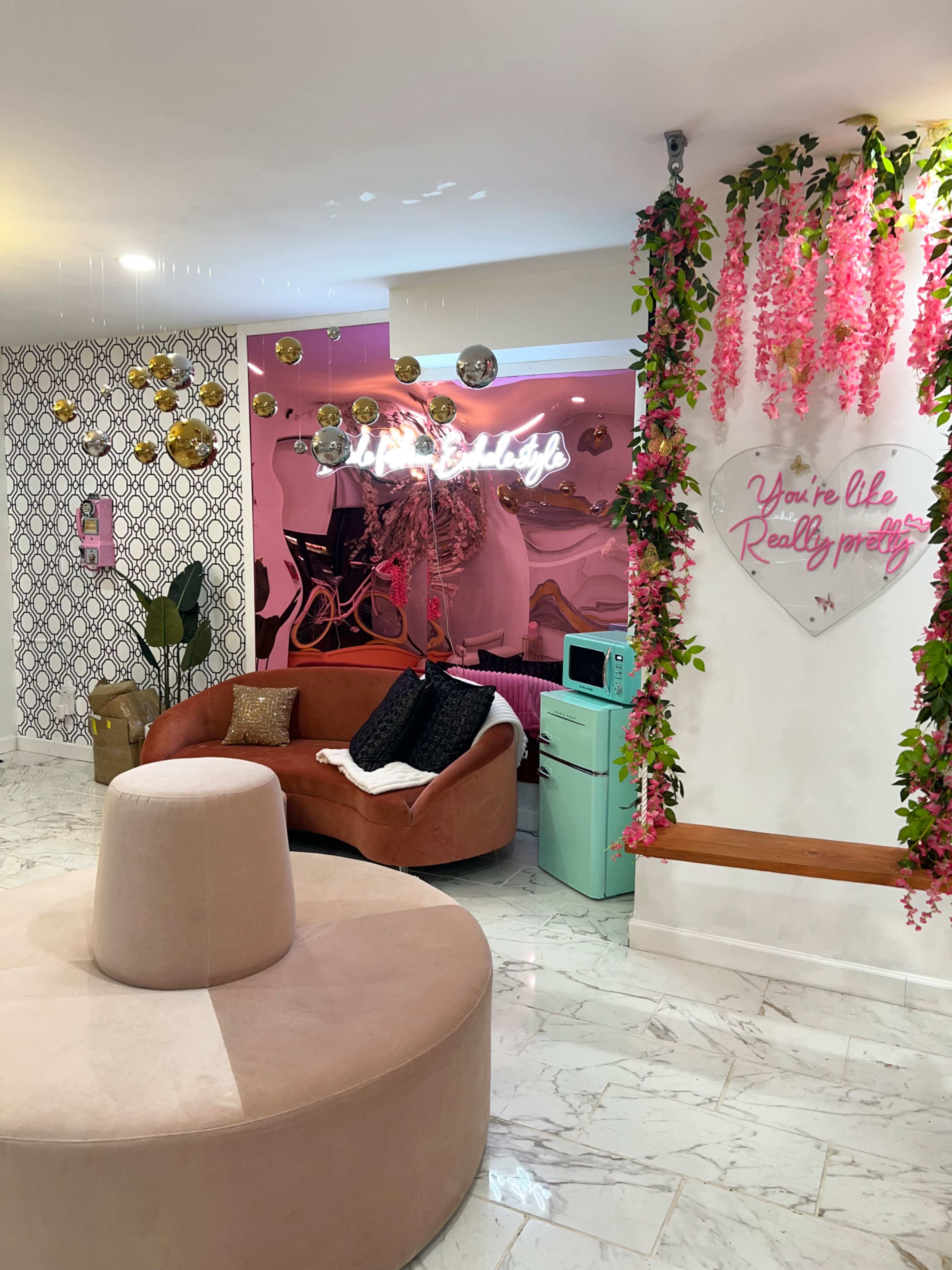 A modern lounge area featuring a circular pink seating arrangement, a curved brown sofa, and walls decorated with patterned wallpaper and vibrant pink flowers.