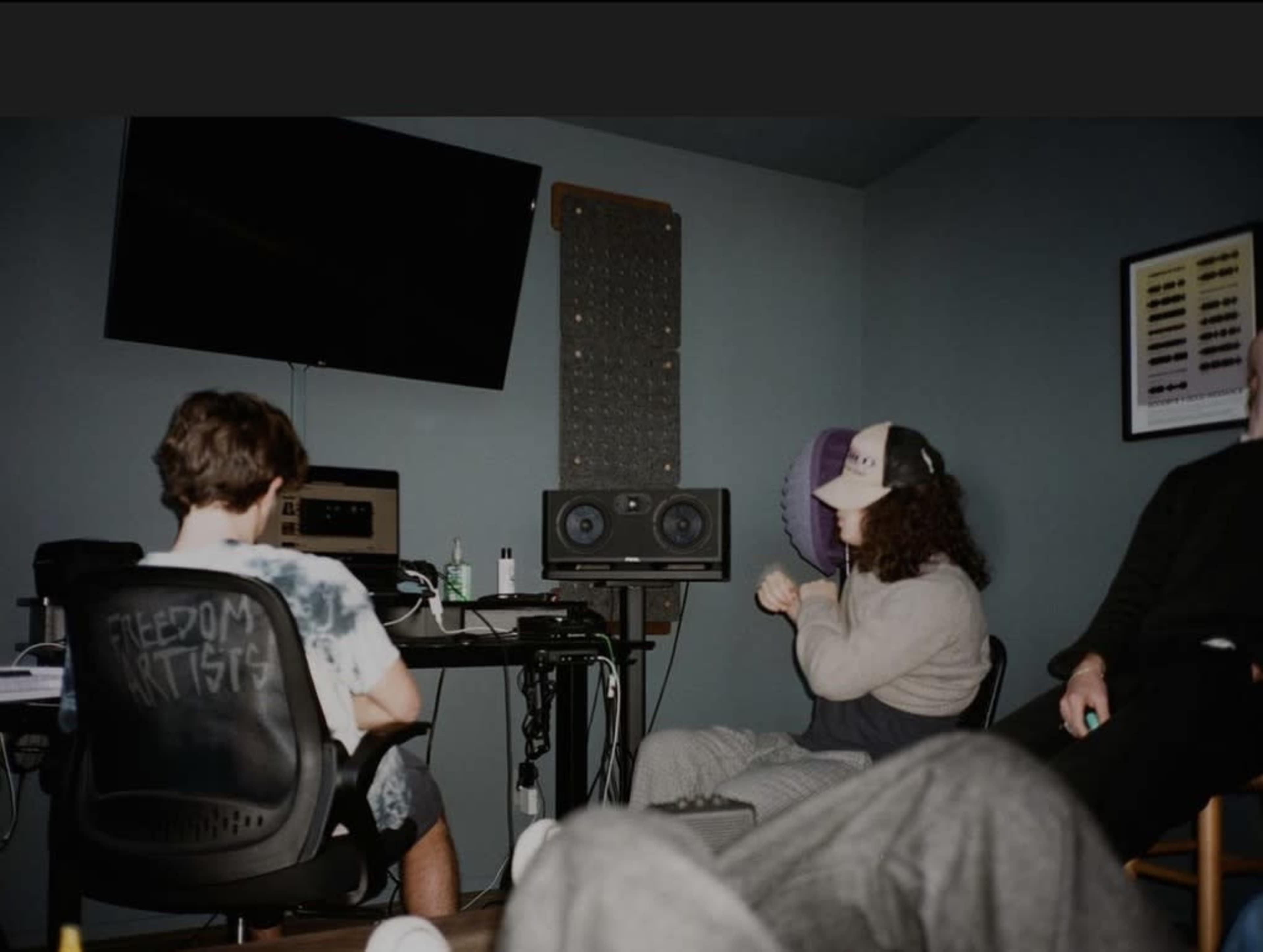 Two individuals sit in a dimly lit room, one working at a computer with audio equipment, while another sits in a chair, engaged in conversation, with a third person partly visible in the foreground.