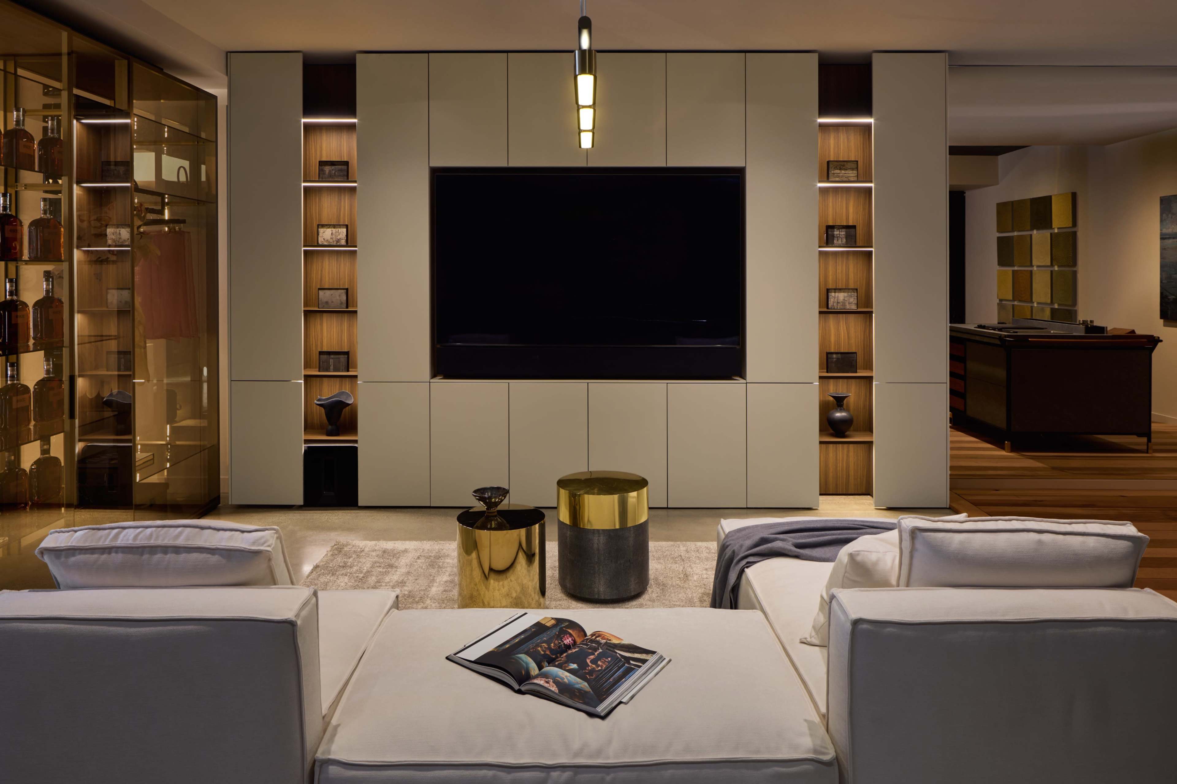 The image shows a modern living room with a large wall-mounted television, sleek cabinetry, and a minimalist seating arrangement featuring white sofas and decorative tables.