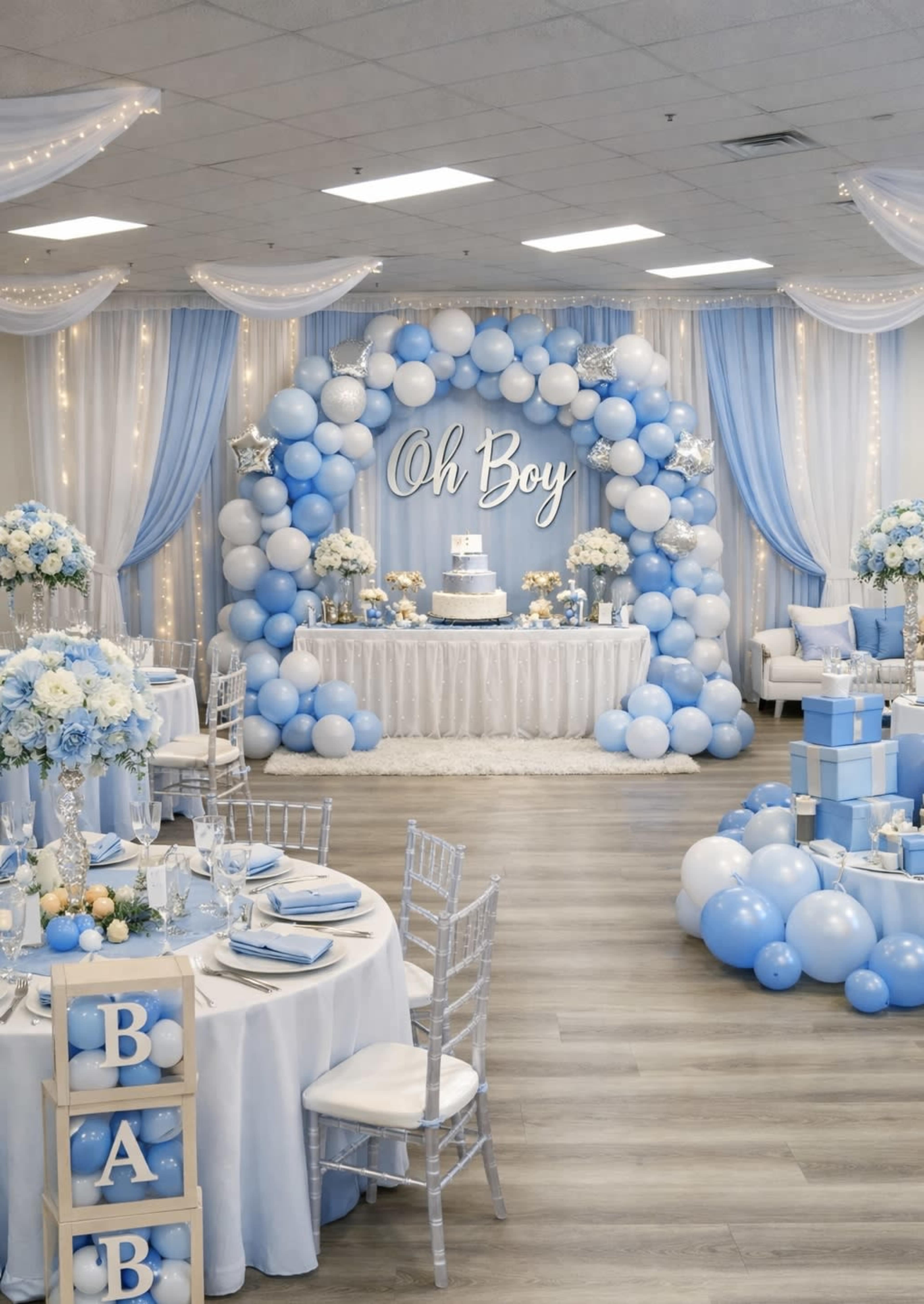 The image shows a decorated event space with blue and white balloons, floral arrangements, and a cake display, set up for a celebration.