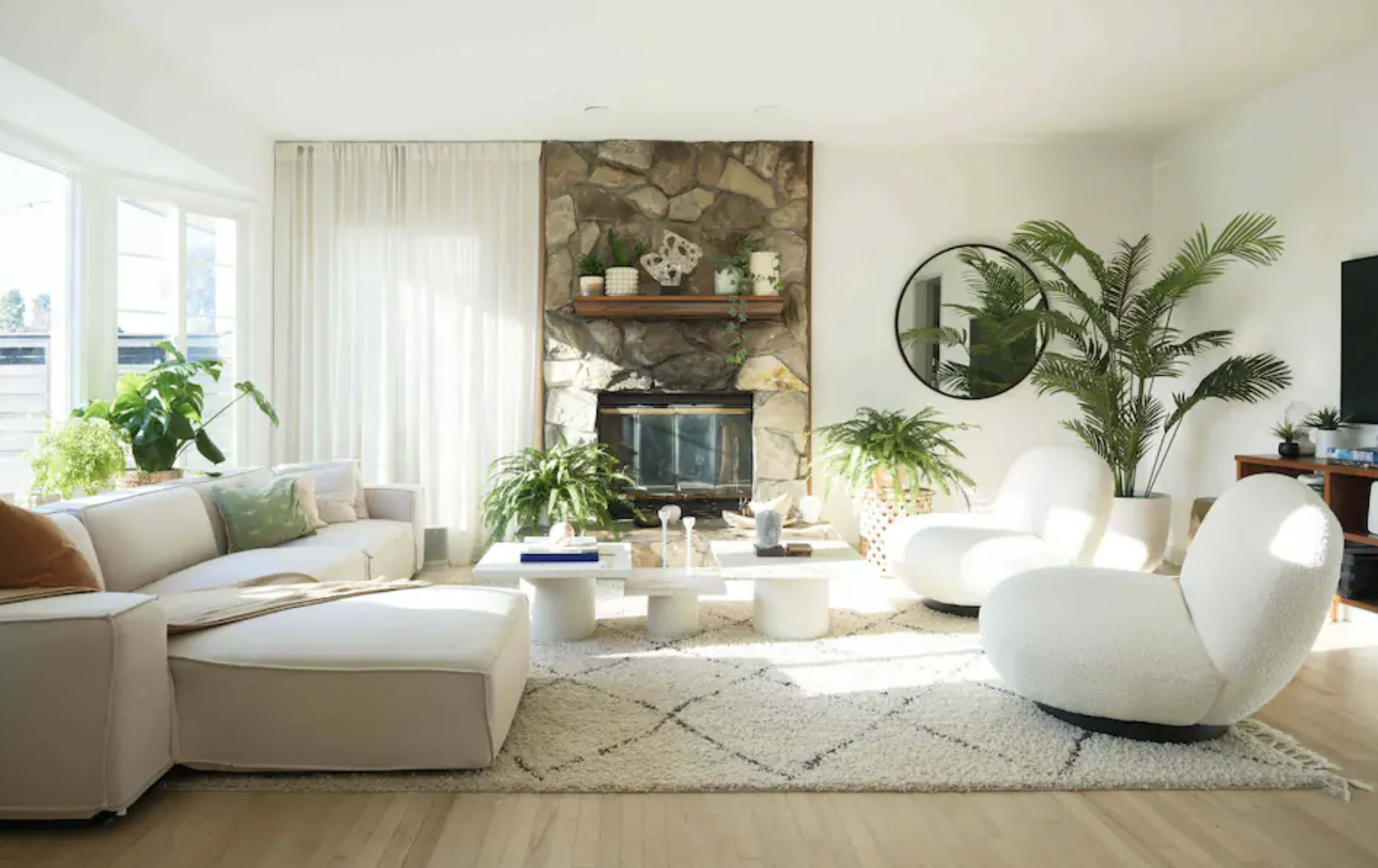 The living room features a large sectional sofa, a stone fireplace with a wooden shelf above, and potted plants scattered throughout the space, all arranged on a cream-colored area rug.