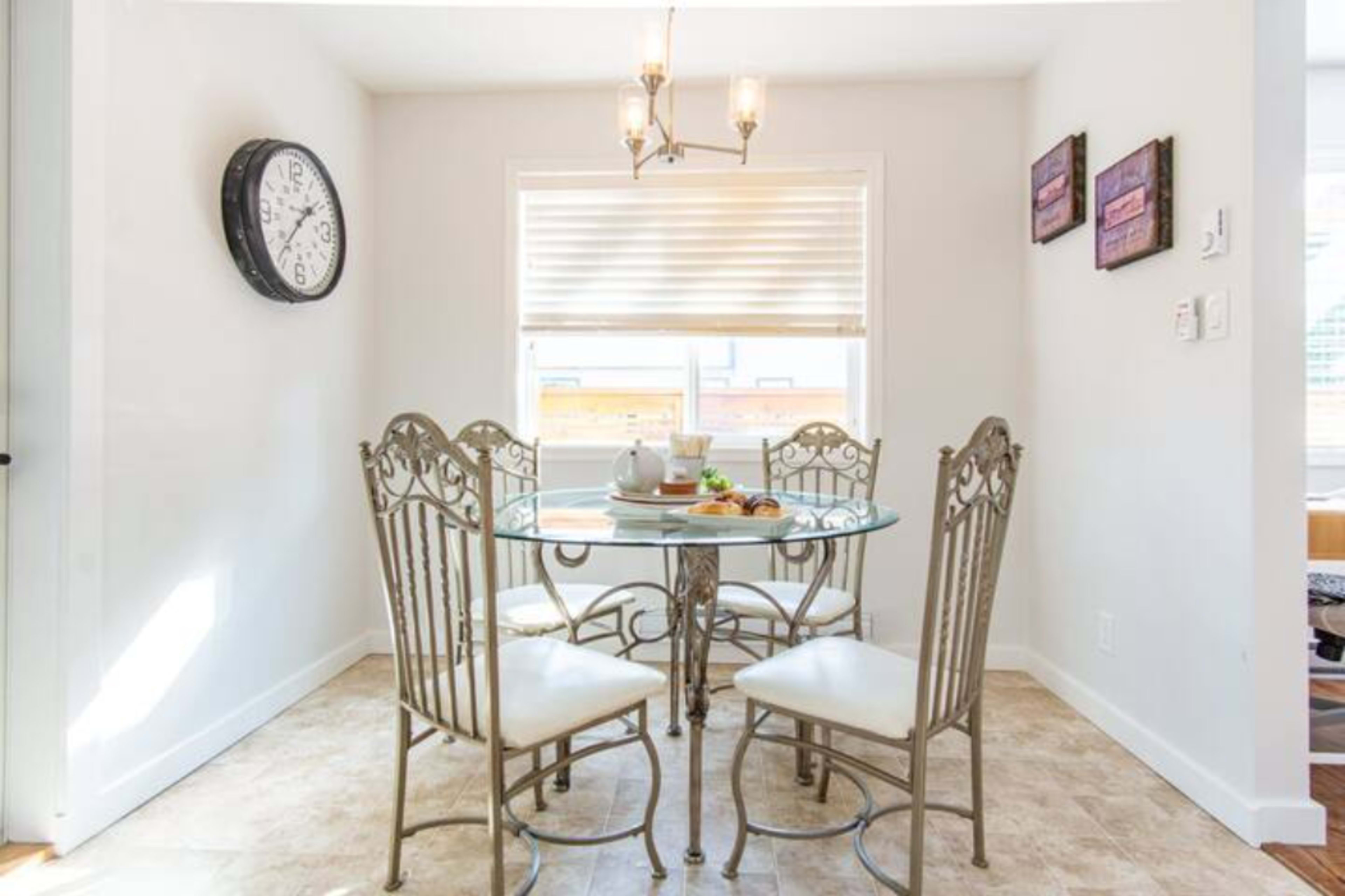 A small dining area features a round glass table with four metal chairs and a clock on the wall, lit by a pendant fixture.