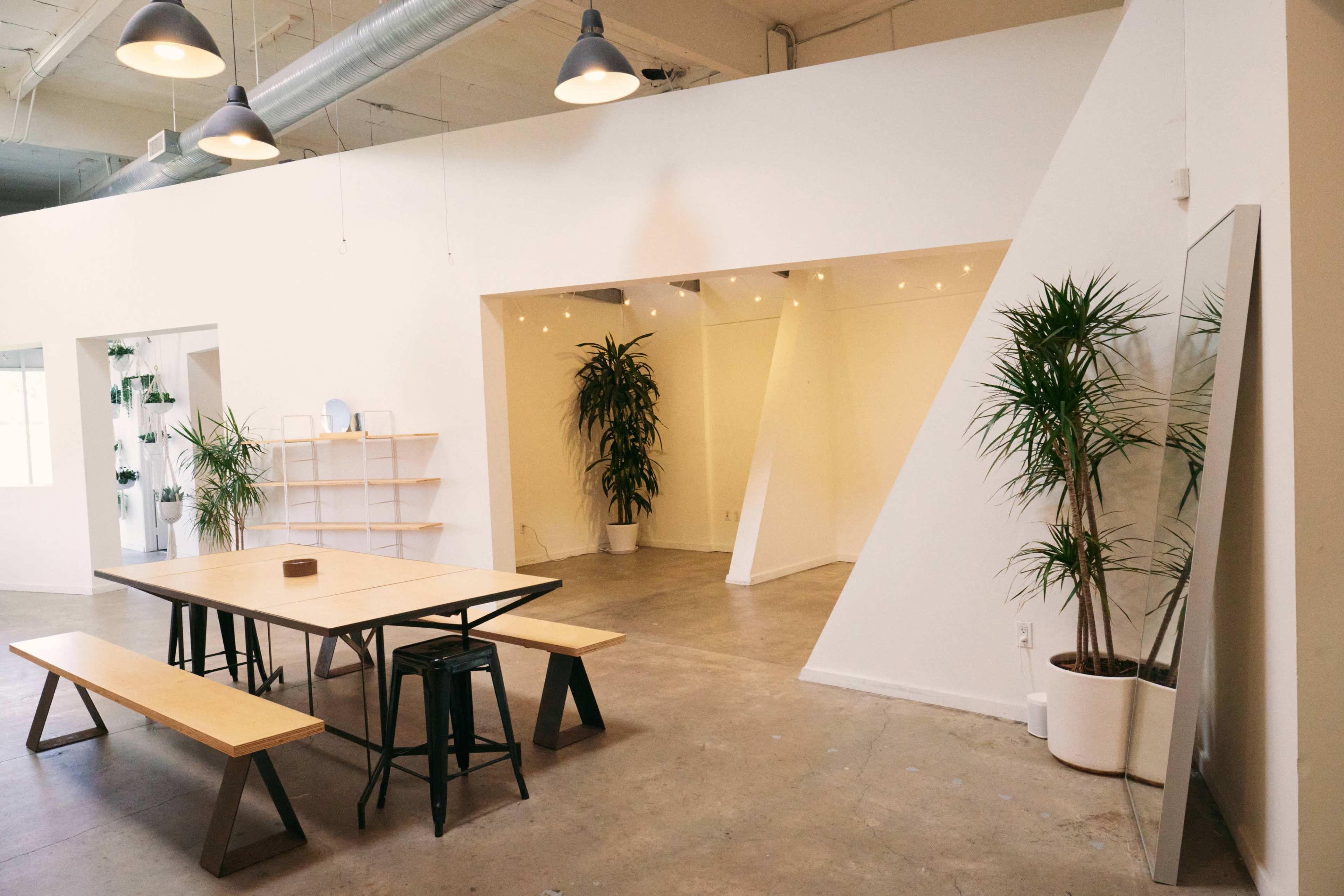 A modern, minimalist interior space featuring a wooden table with benches, surrounded by potted plants and a large mirror against a white wall.