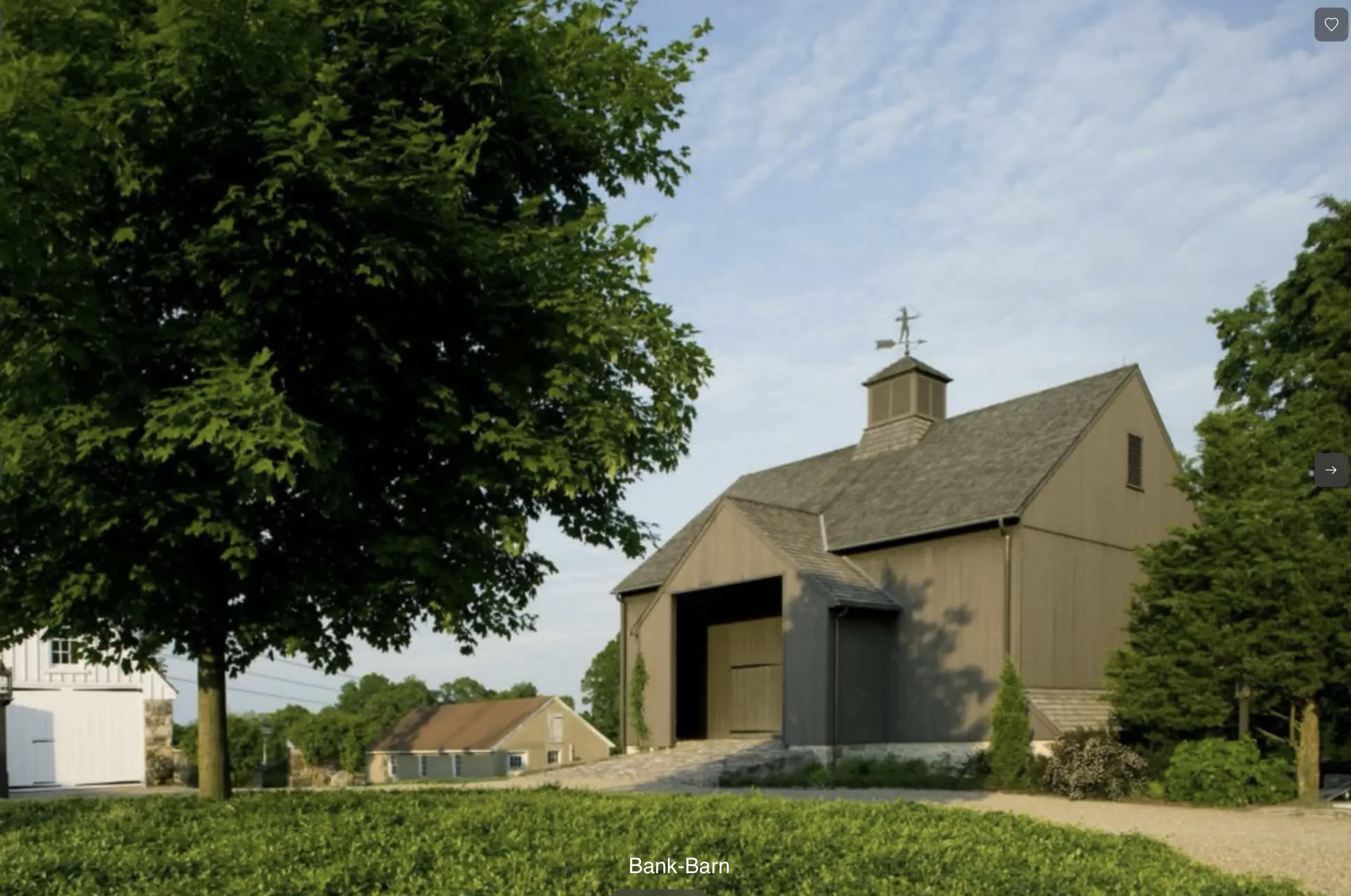 The image shows a bank barn surrounded by greenery and a large tree, with a clear sky in the background.