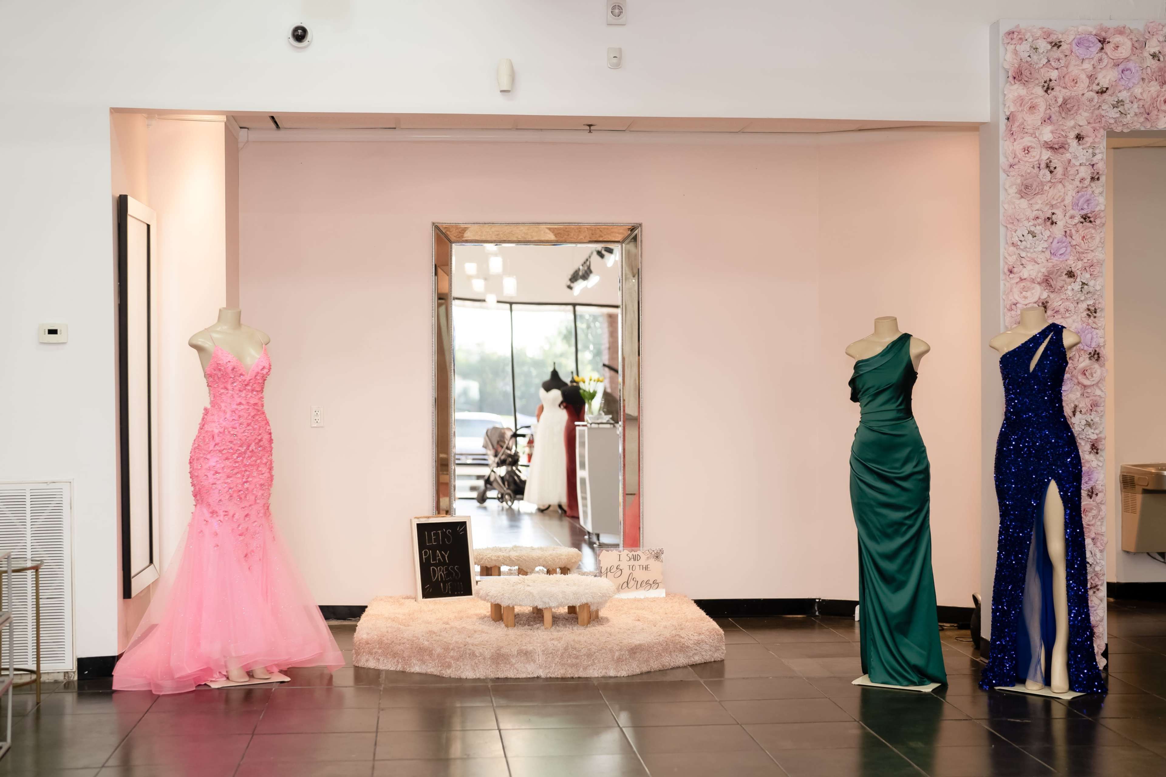 A boutique display featuring three evening gowns on dress forms, with a large mirror and a decorative sign in a stylish interior setting.