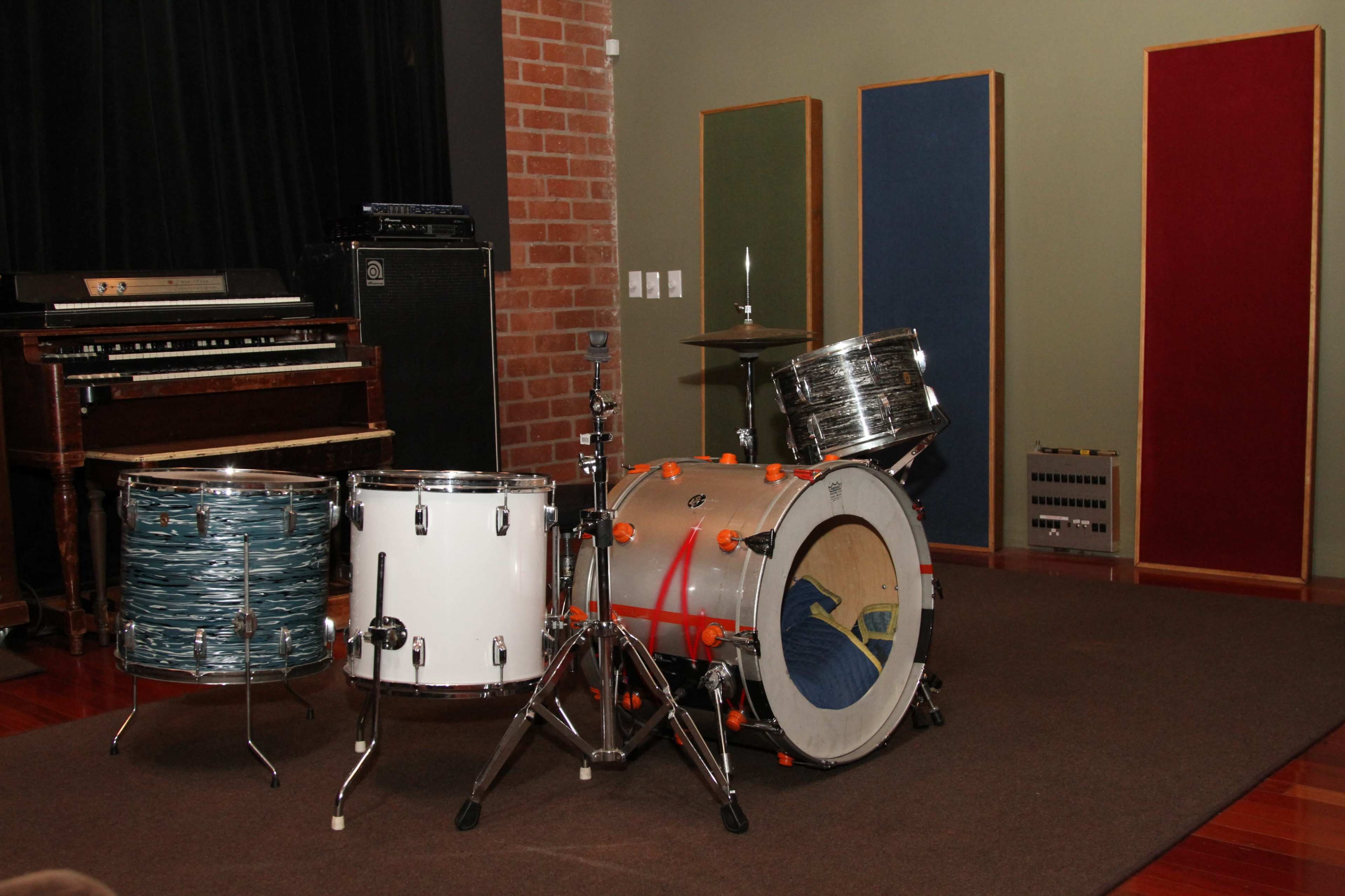 The image features a small drum set consisting of a bass drum, snare drum, and two toms, situated in a music studio with a brick wall and soundproofing panels in the background.