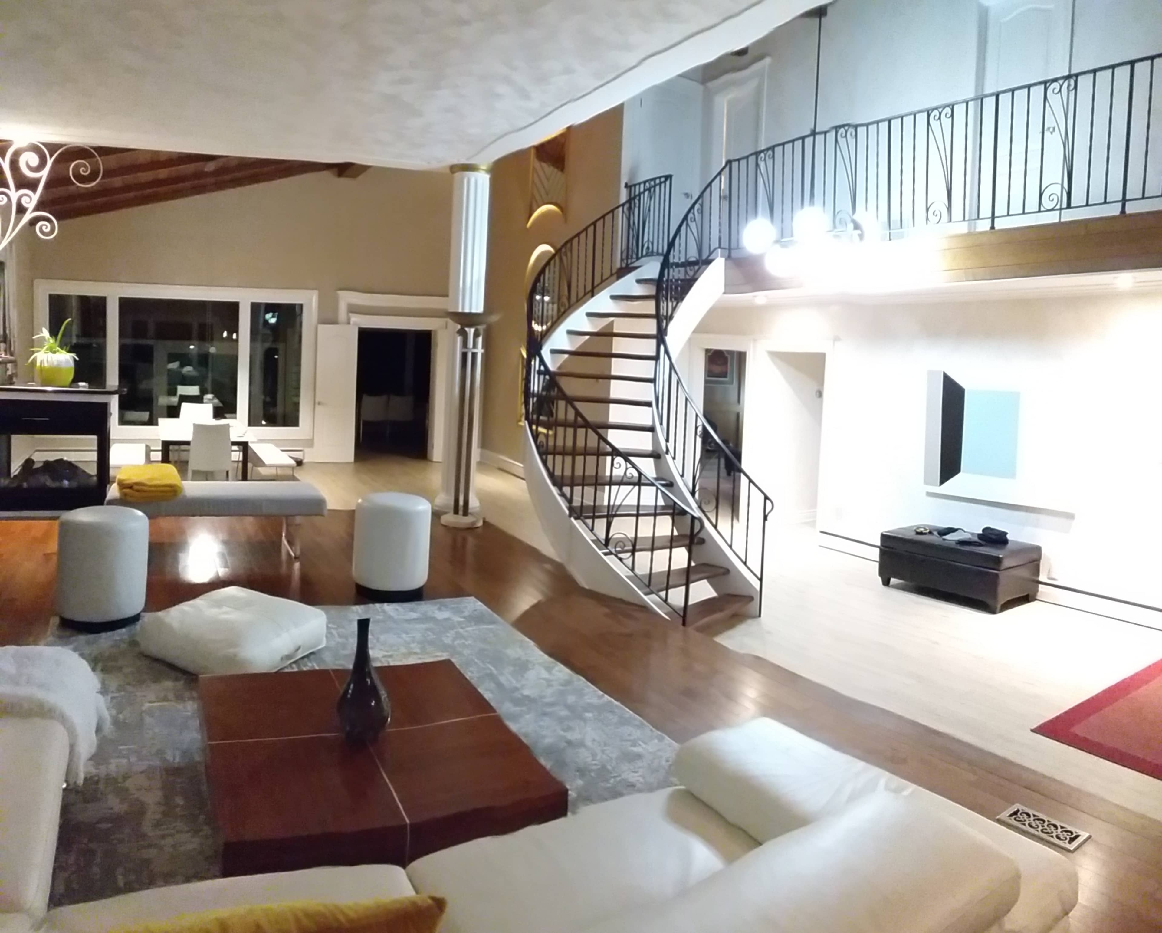 A spacious living room with a modern design, featuring a large staircase, a seating area with a white sofa, and large windows.