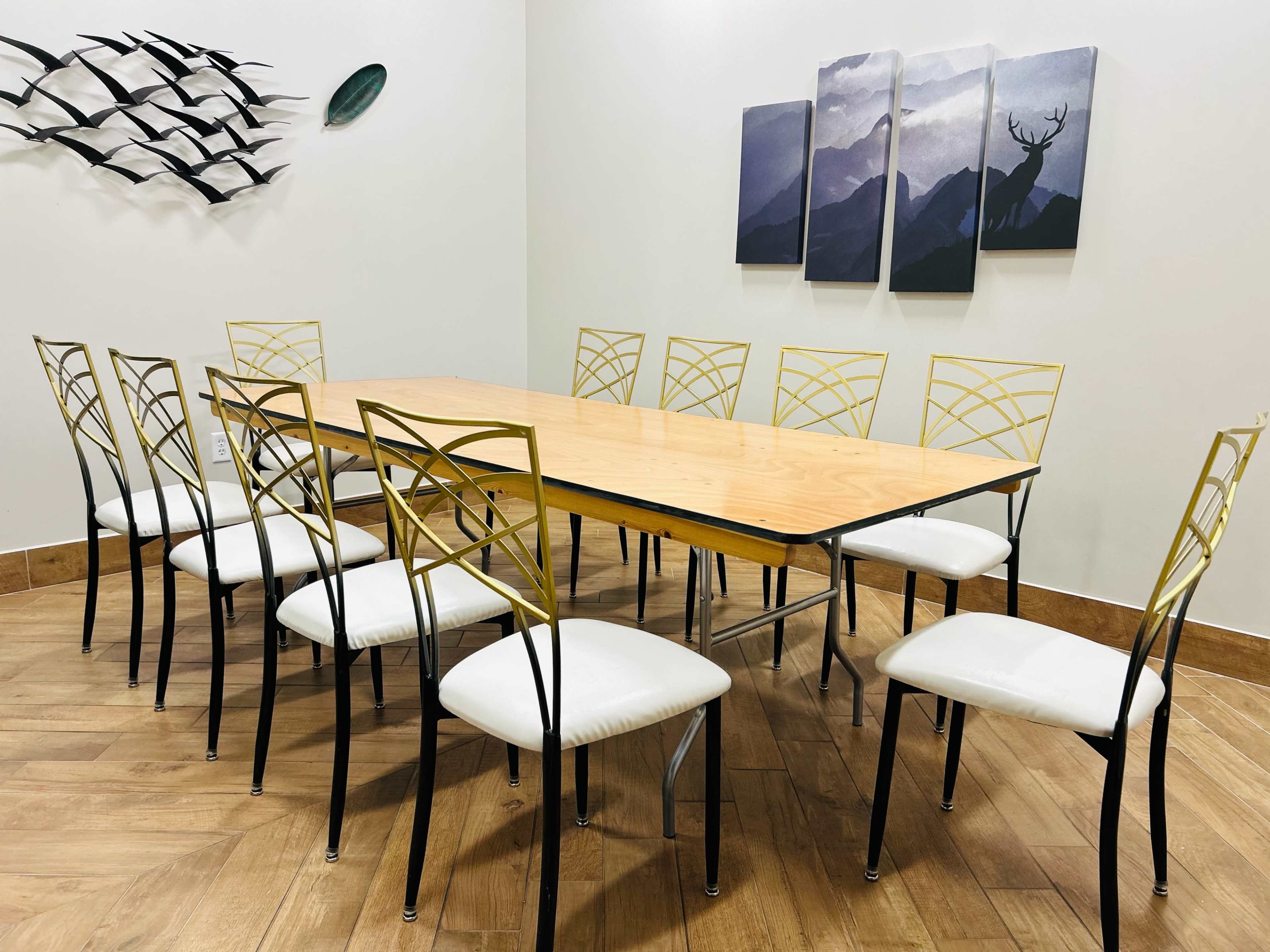 A wooden table surrounded by twelve metal and white upholstered chairs is placed in a room with mounted artwork depicting mountains and a deer.