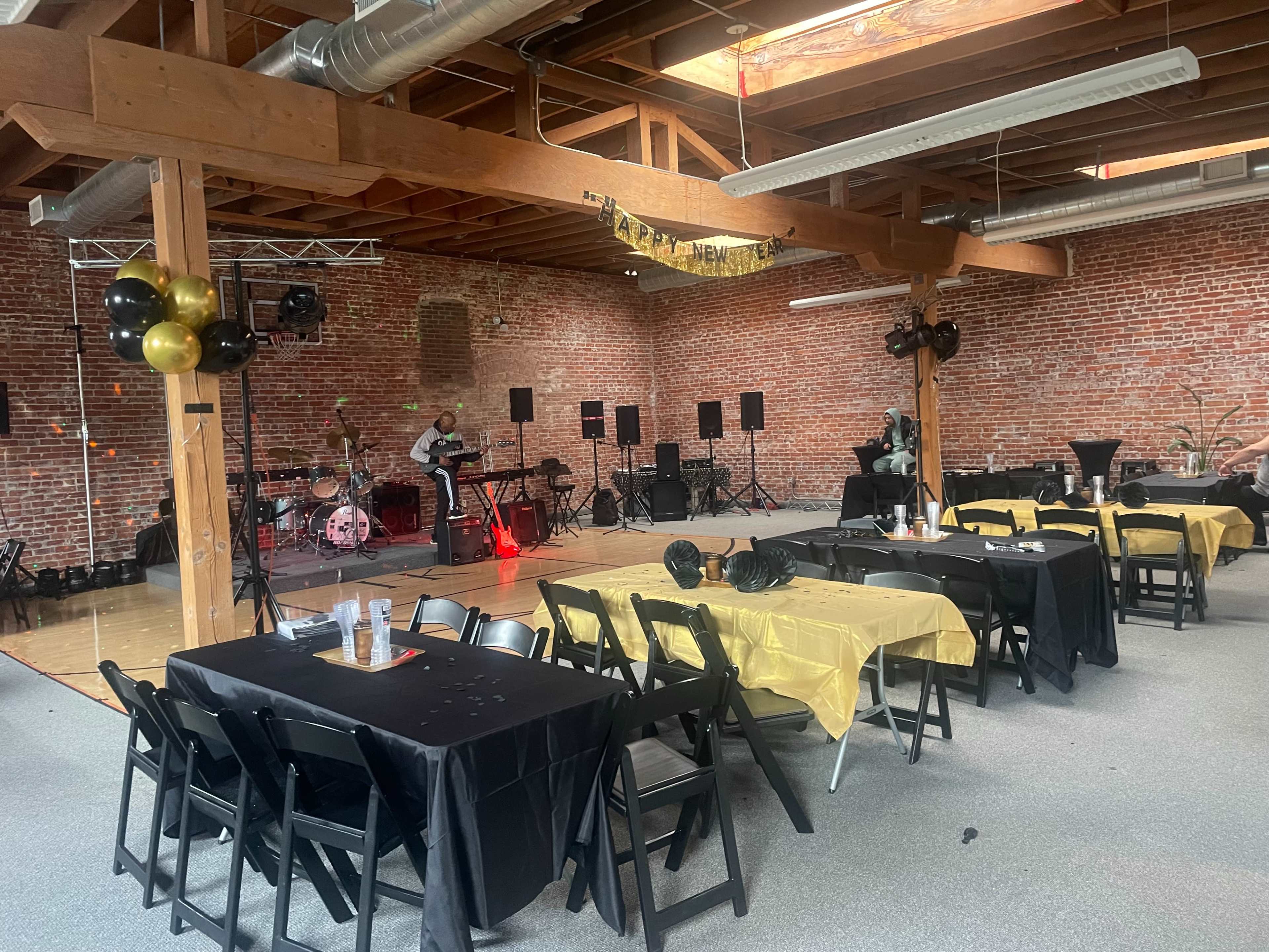 The image shows a spacious indoor venue with brick walls, several tables decorated in black and gold, and a stage set up for a live performance.