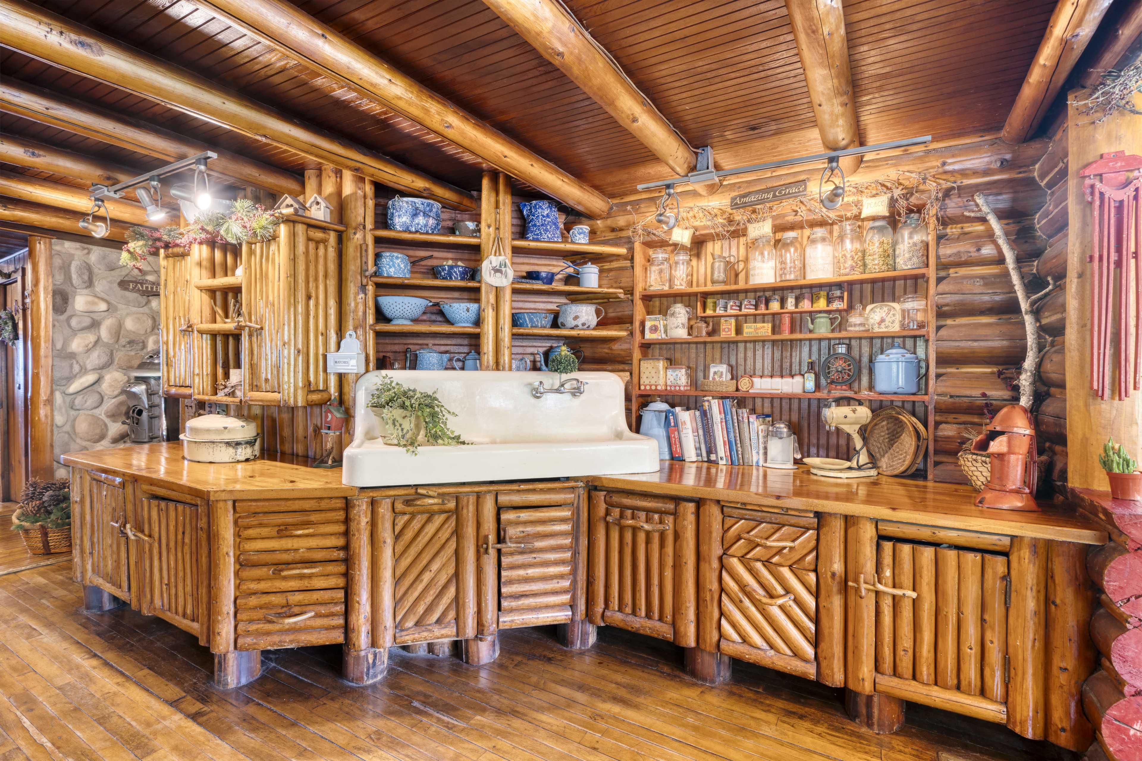 The image shows a rustic kitchen with wooden cabinets, a farmhouse sink, and shelves filled with dishes and decorative items.