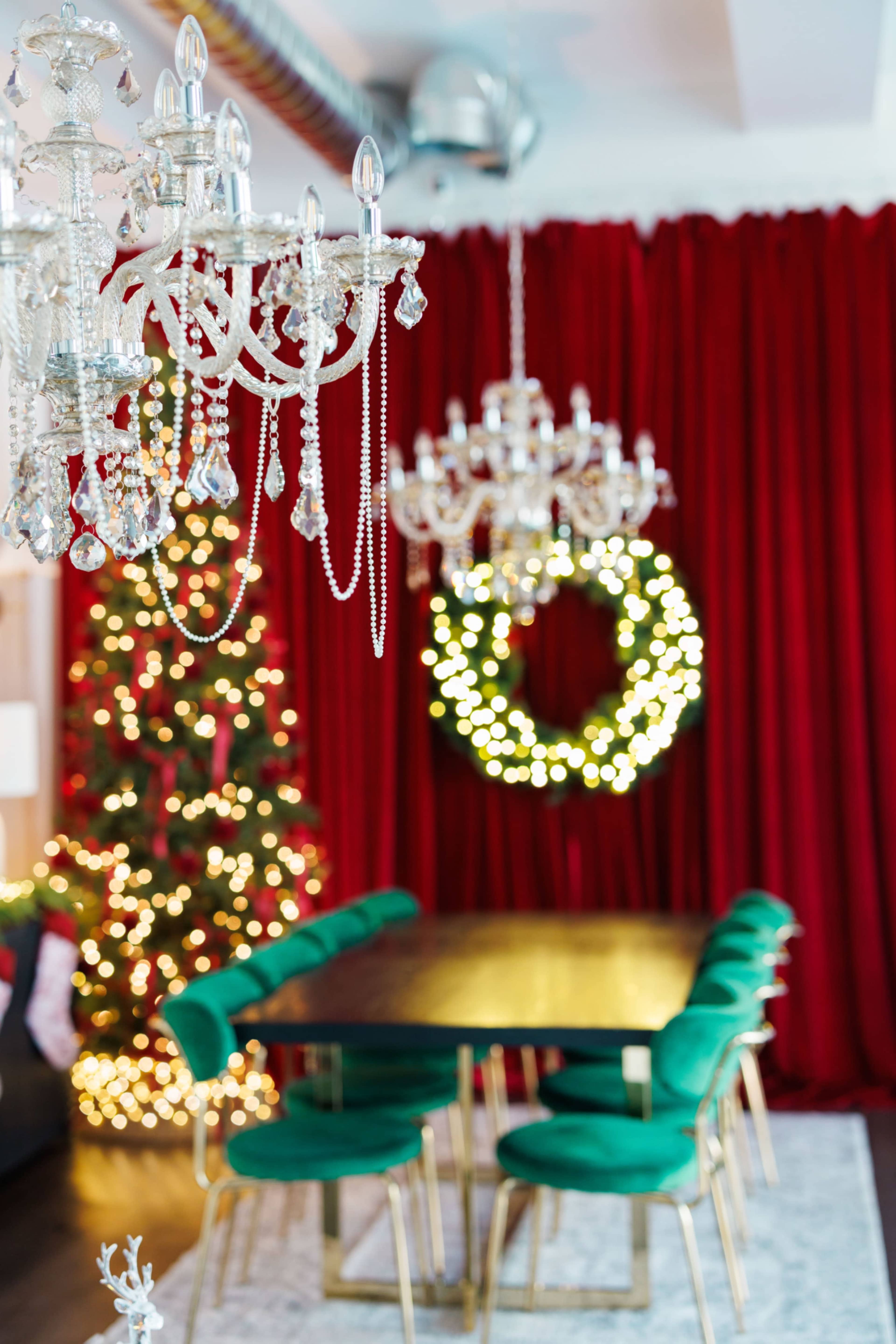 A festive dining area features a dark table surrounded by green chairs, with a decorated Christmas tree and a lit wreath in the background, all complemented by ornate chandeliers.