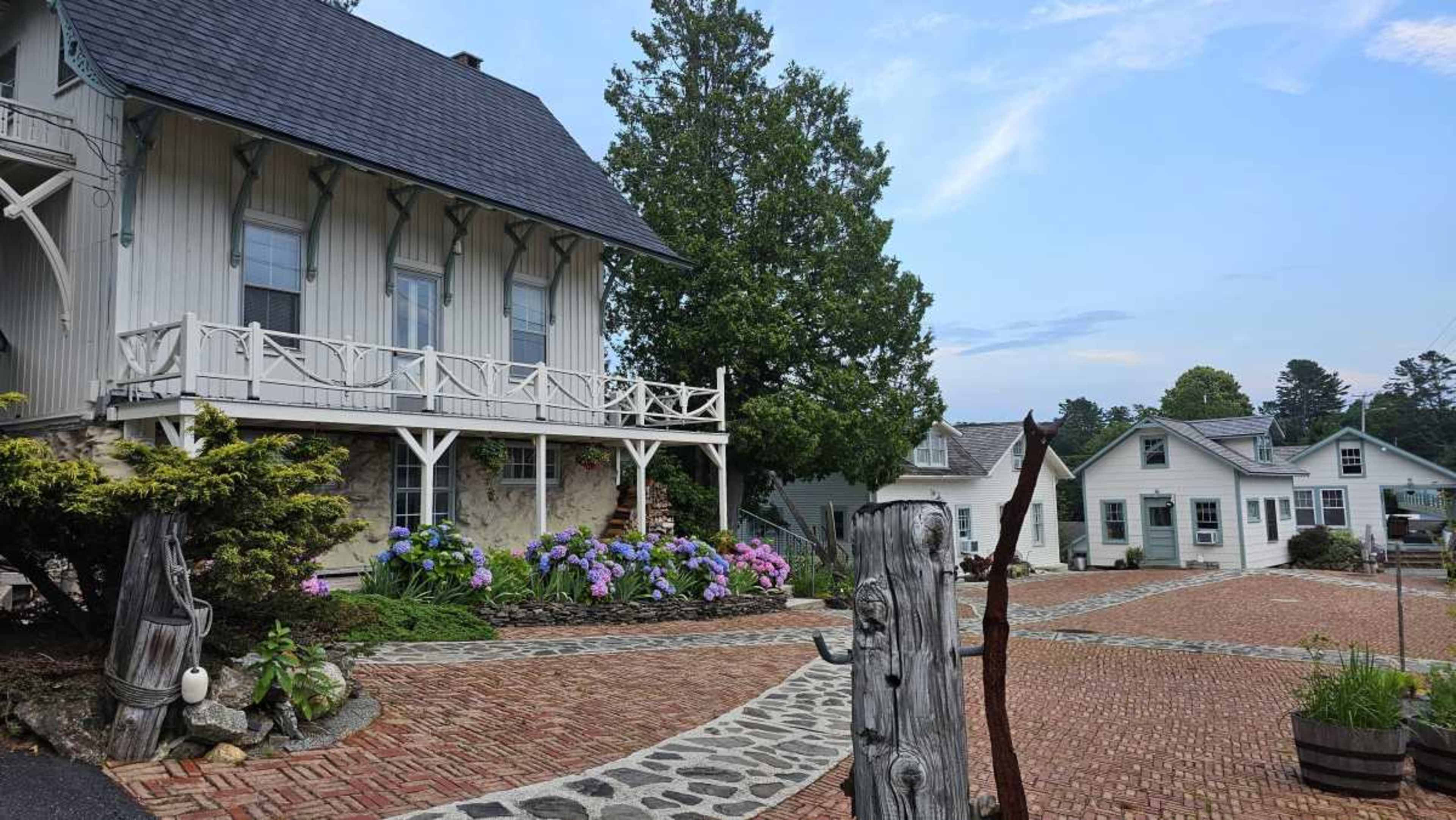 The image shows a cluster of quaint buildings surrounded by blooming flowers and a stone pathway.