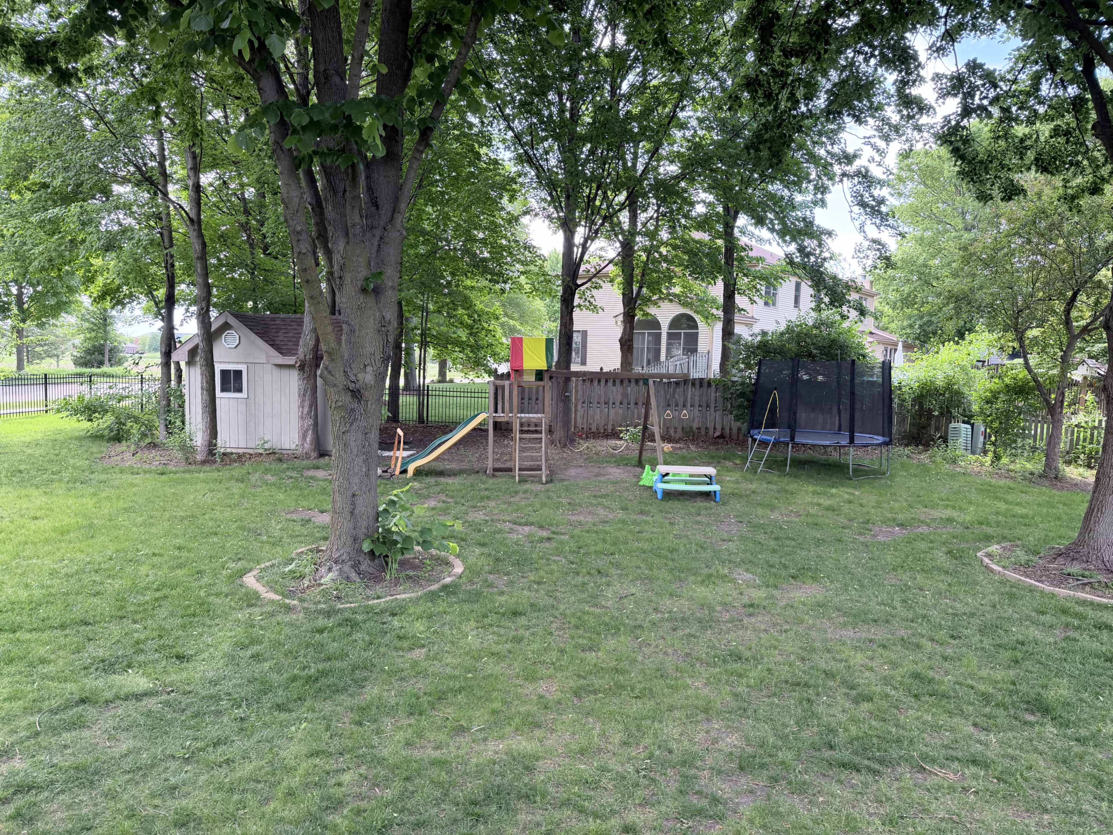 The image shows a backyard featuring a playground with a slide, a wooden playhouse, a trampoline, and green grass surrounded by trees.