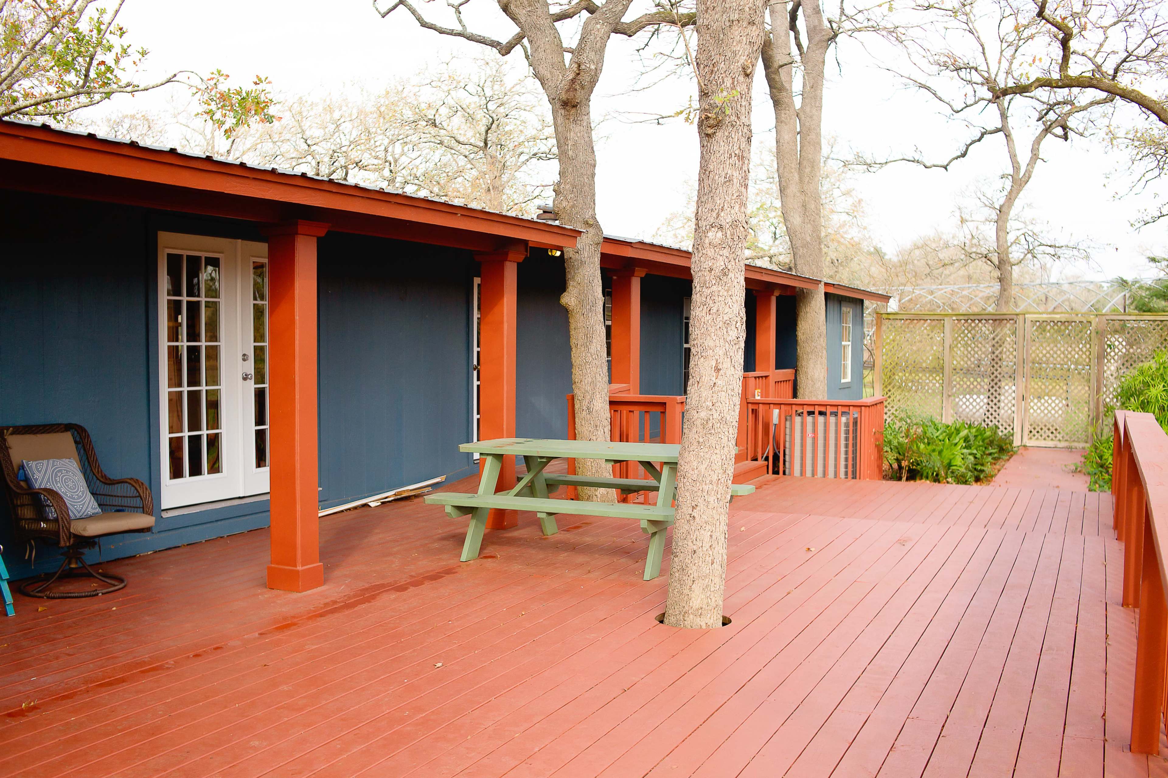 A wooden deck with a green picnic table is situated in front of a building featuring a blue exterior and white French doors.