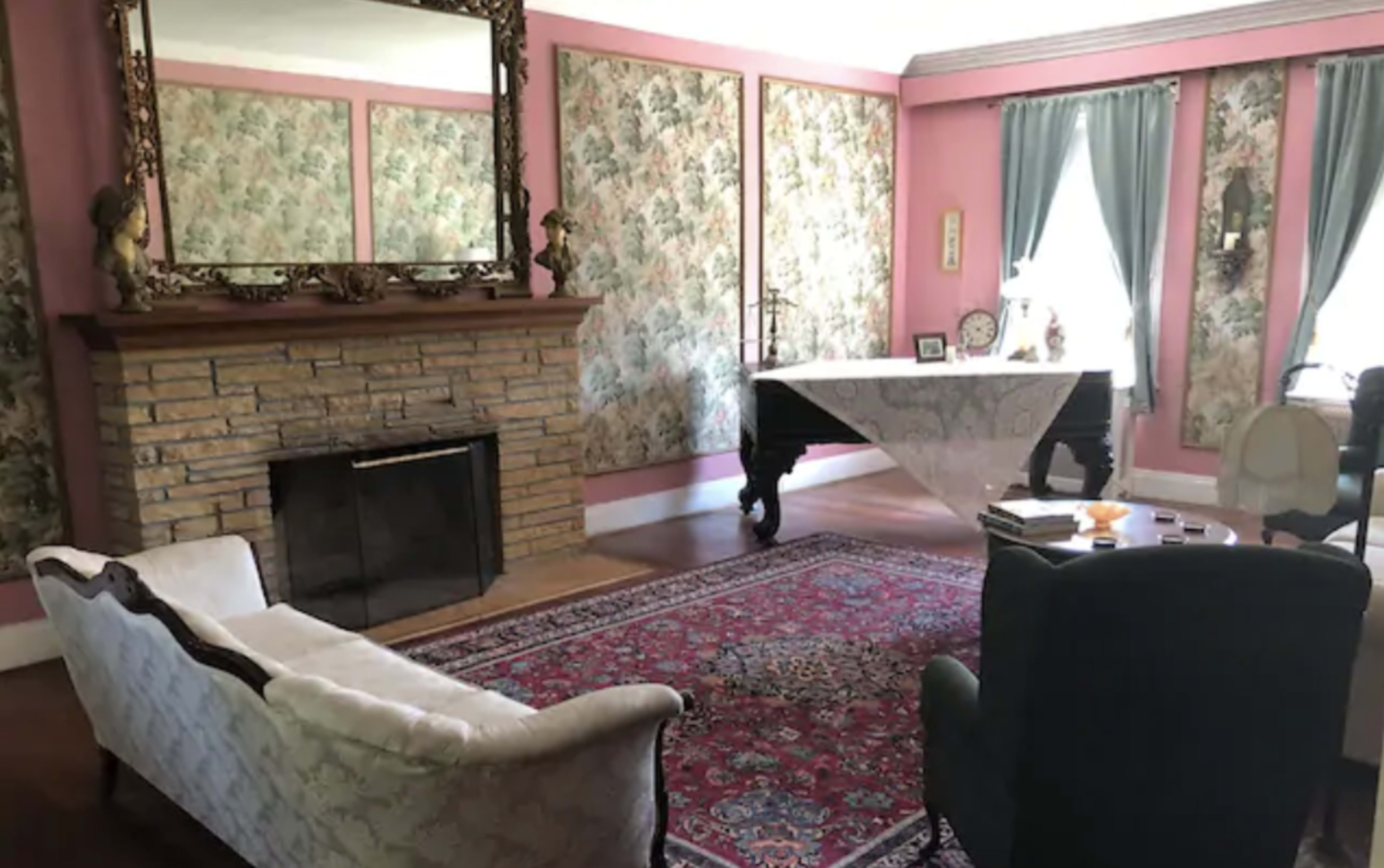 The room features a patterned wallpaper, a stone fireplace, a large table covered with a lace tablecloth, and a mix of vintage furniture including a sofa and armchair.