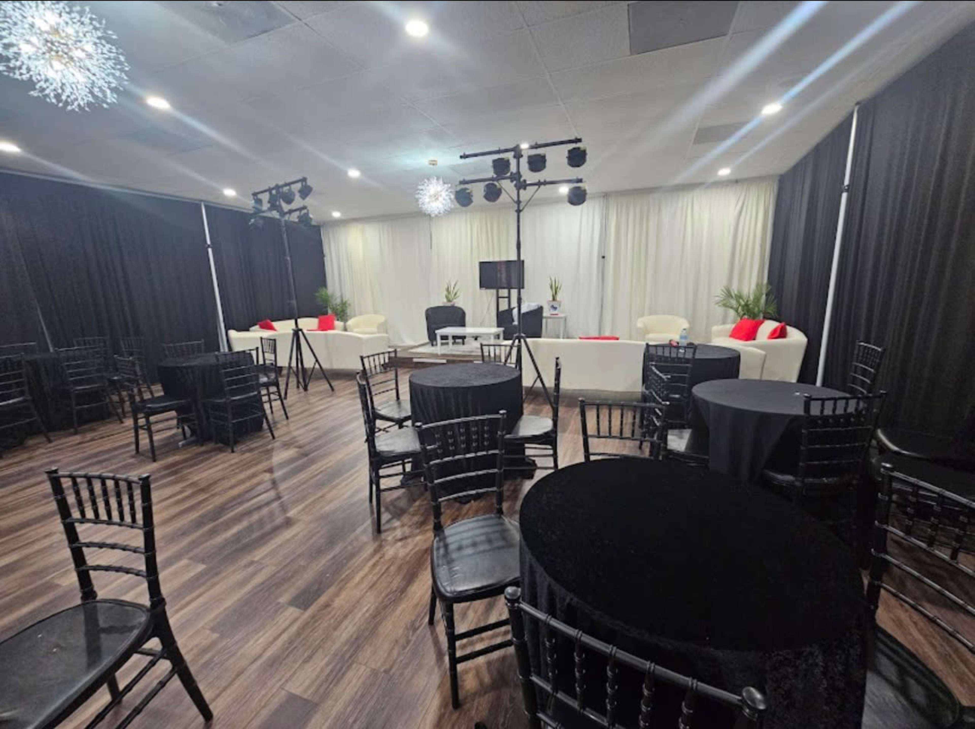 The image shows a large indoor event space with black tables and chairs arranged around the room, featuring a stage area with white seating and decorative lighting.