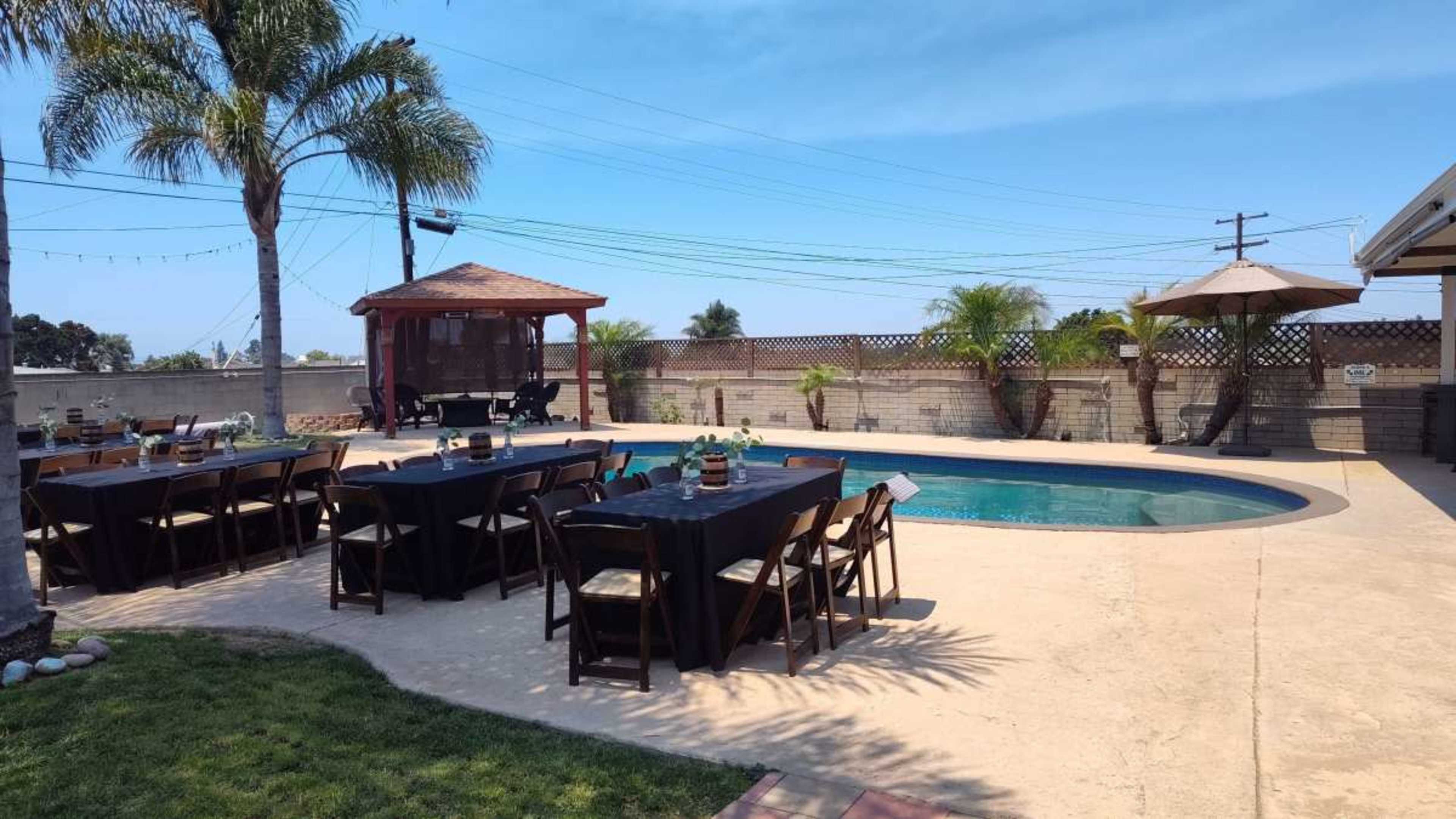 The image shows an outdoor space with a swimming pool, surrounded by tables set for dining, palm trees, and a gazebo in the background.