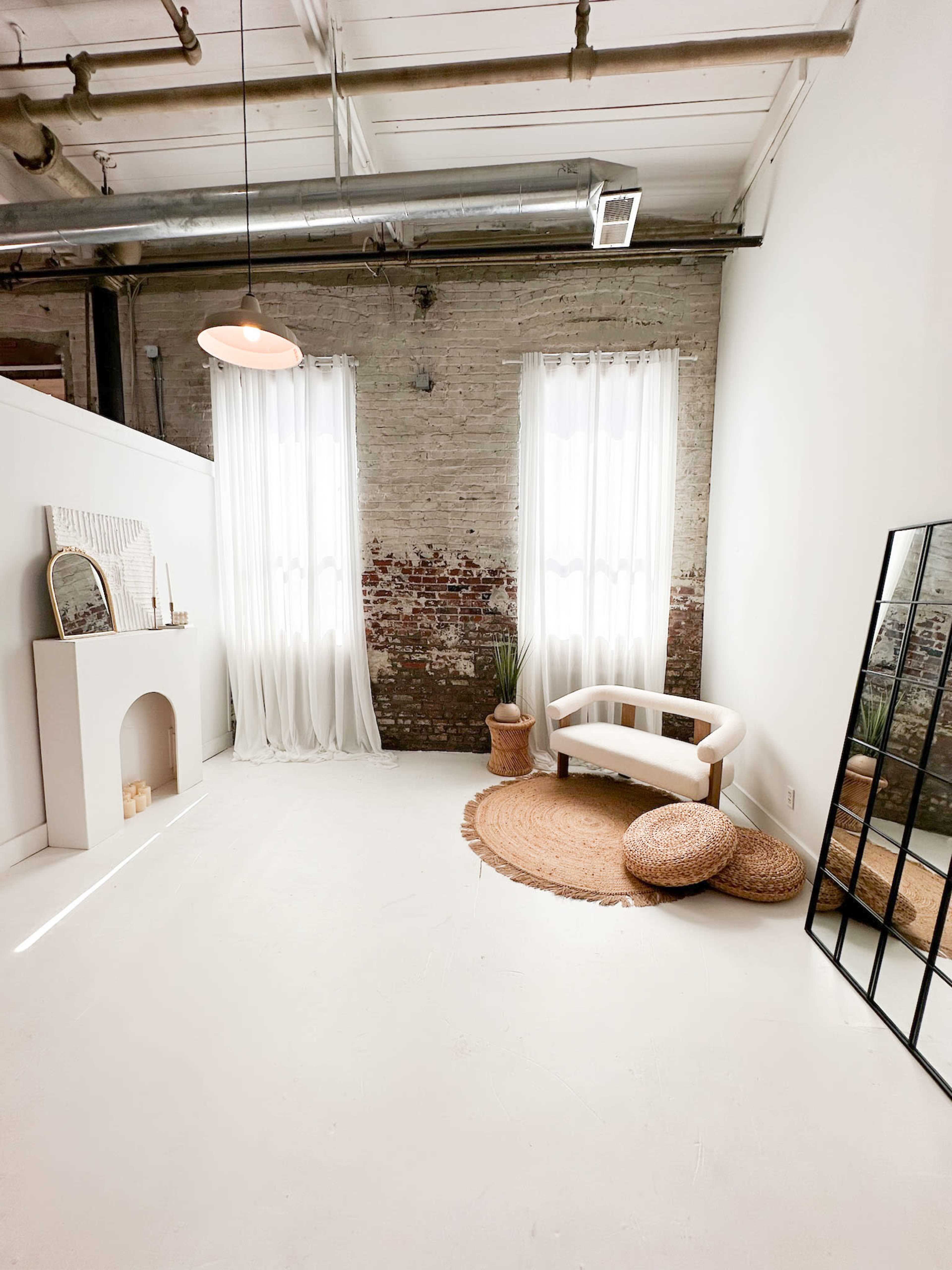 The scene features a bright, minimalist room with exposed brick walls, two large windows dressed in sheer curtains, a white sofa, a round rug, and a full-length mirror.