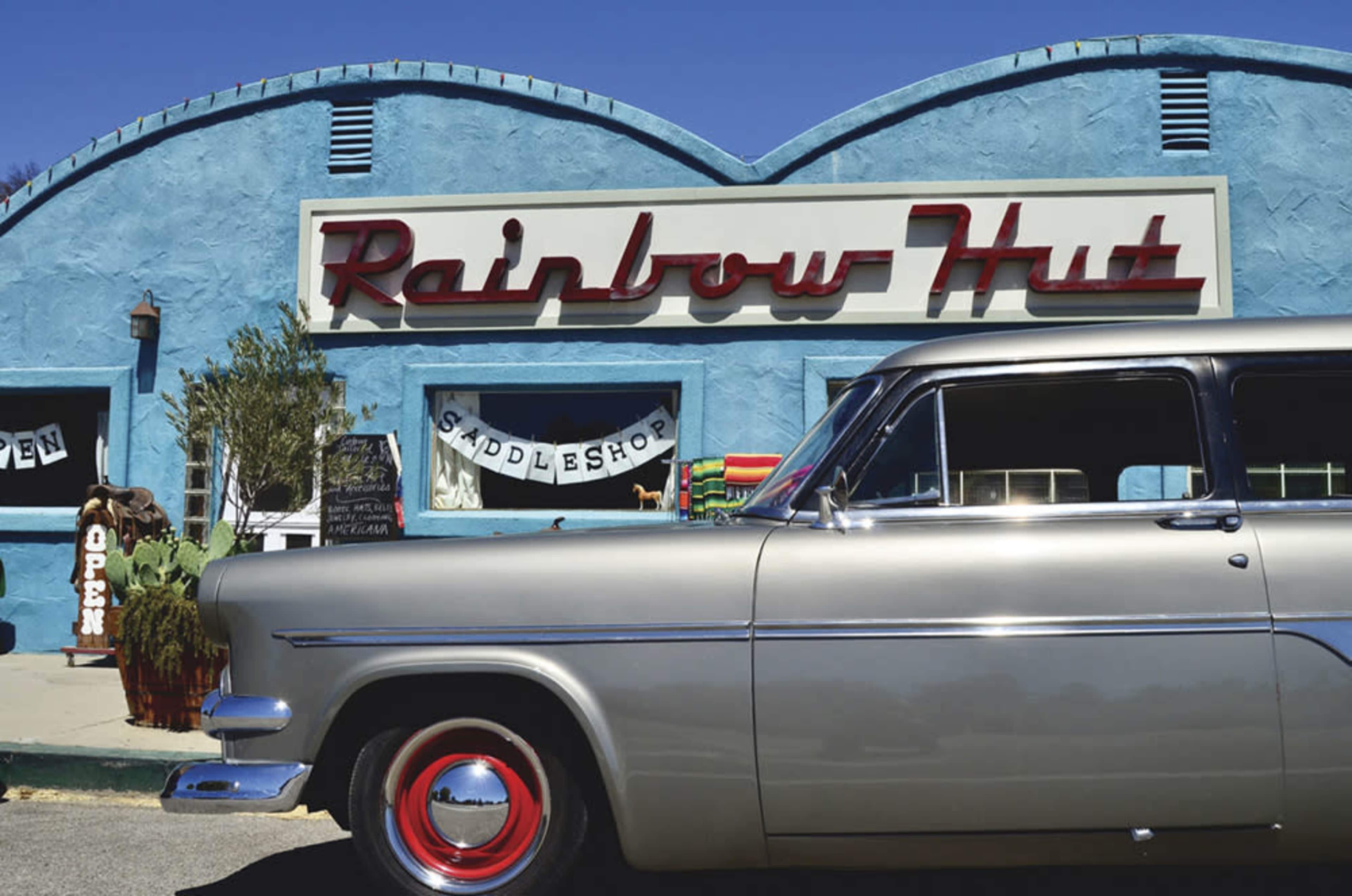 A classic silver car is parked in front of a blue building with the sign "Rainbow Hut Candle Shop."