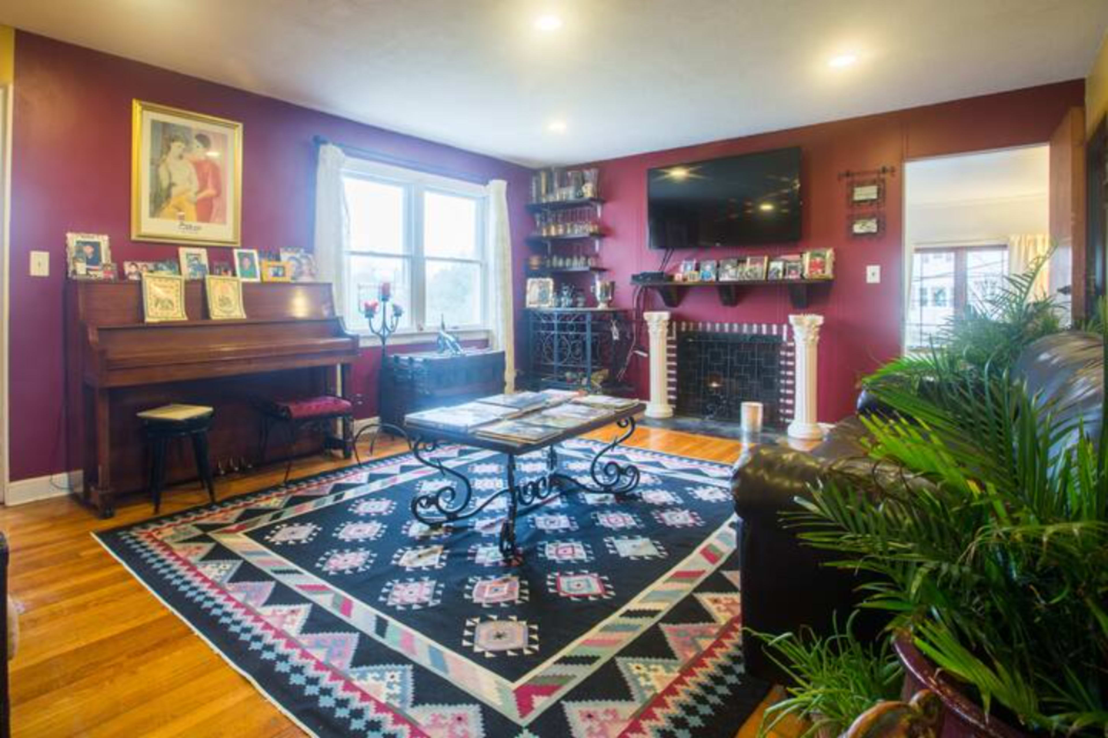 The image shows a living room with a piano, a fireplace, a coffee table, and a large patterned rug, surrounded by framed photographs on shelves and walls.