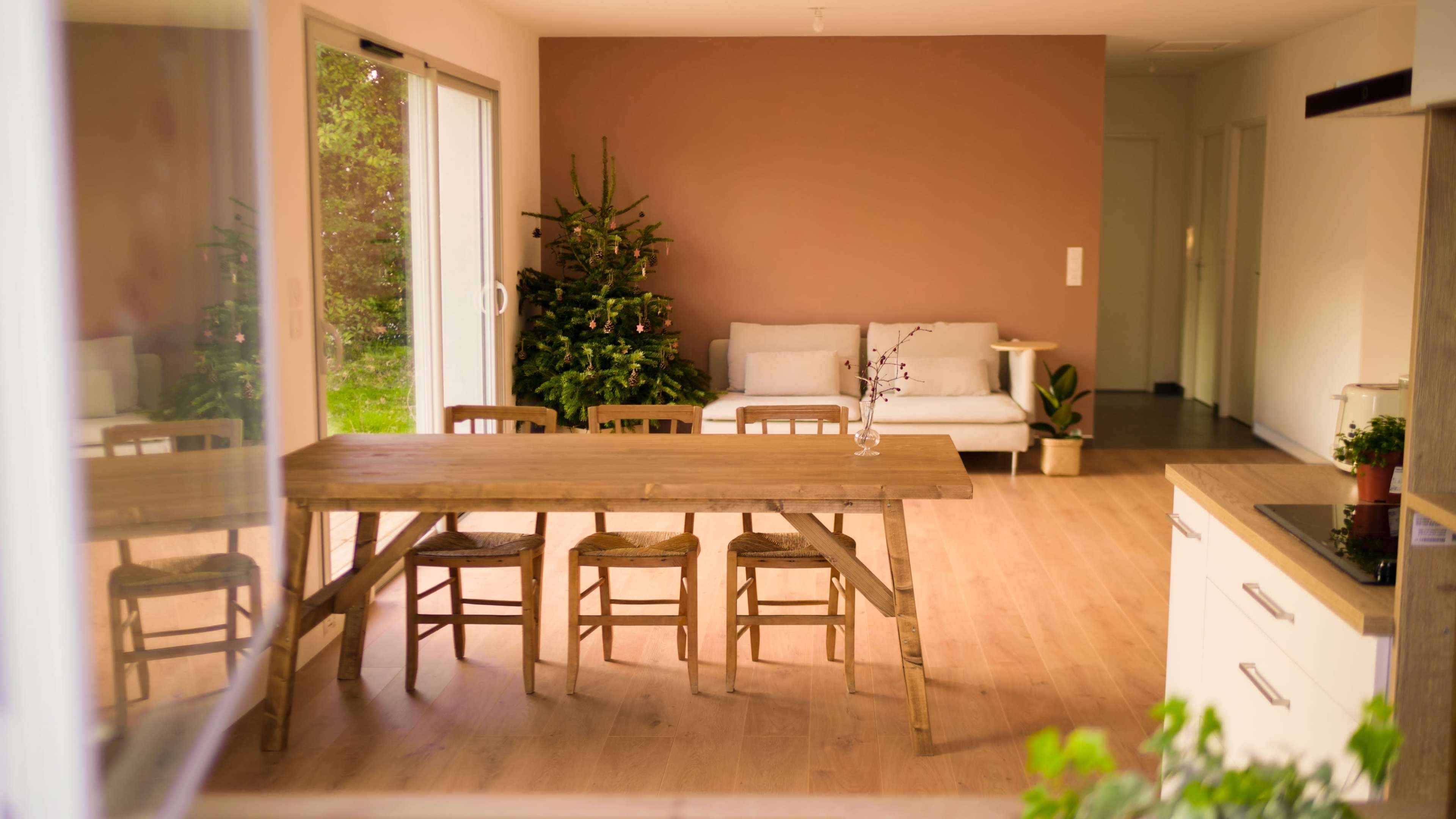 A wooden table with four chairs is set in a bright room featuring a couch, a small plant, and a Christmas tree in the background.