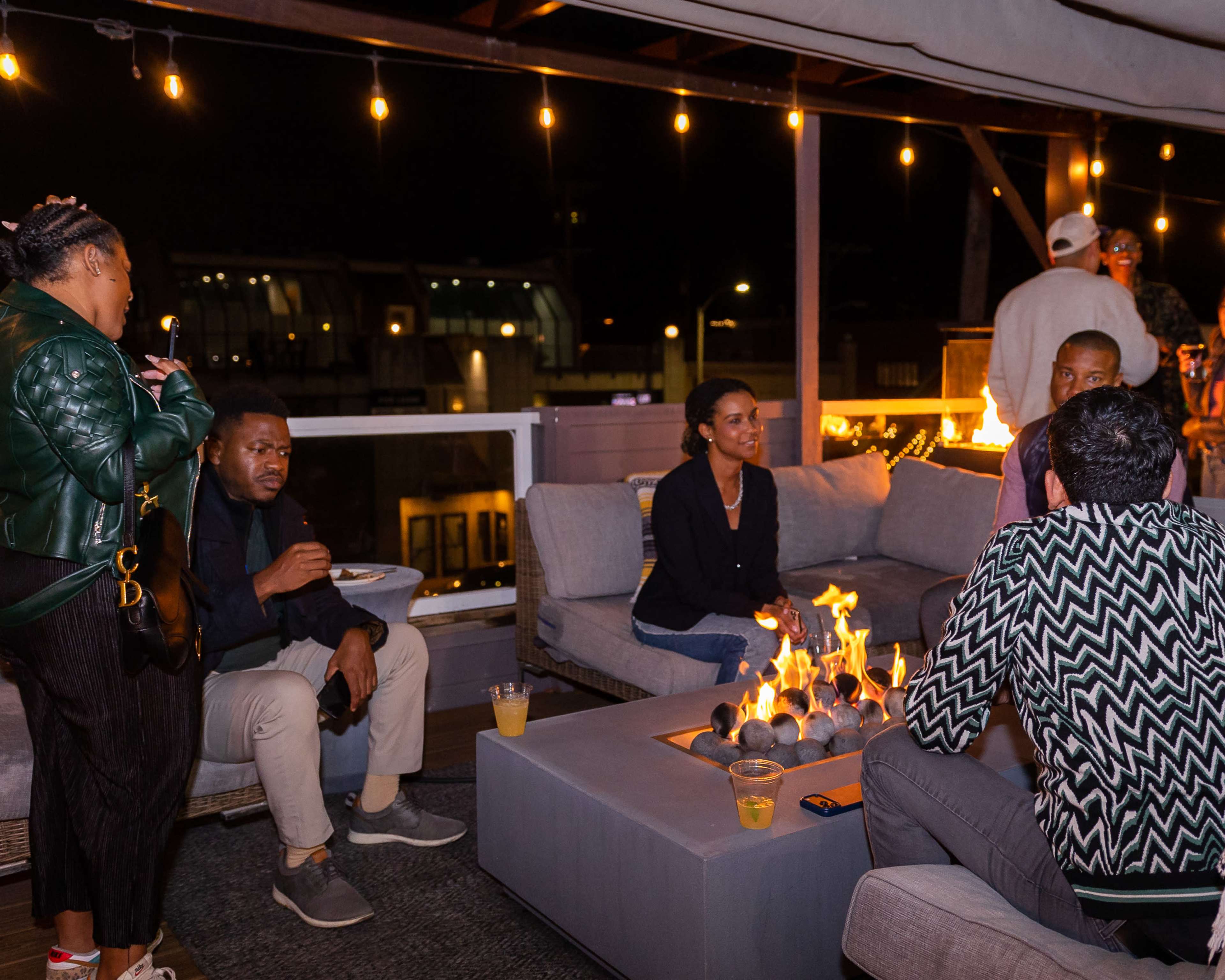 A group of people are seated on a rooftop lounge, socializing around a fire pit while string lights illuminate the night.