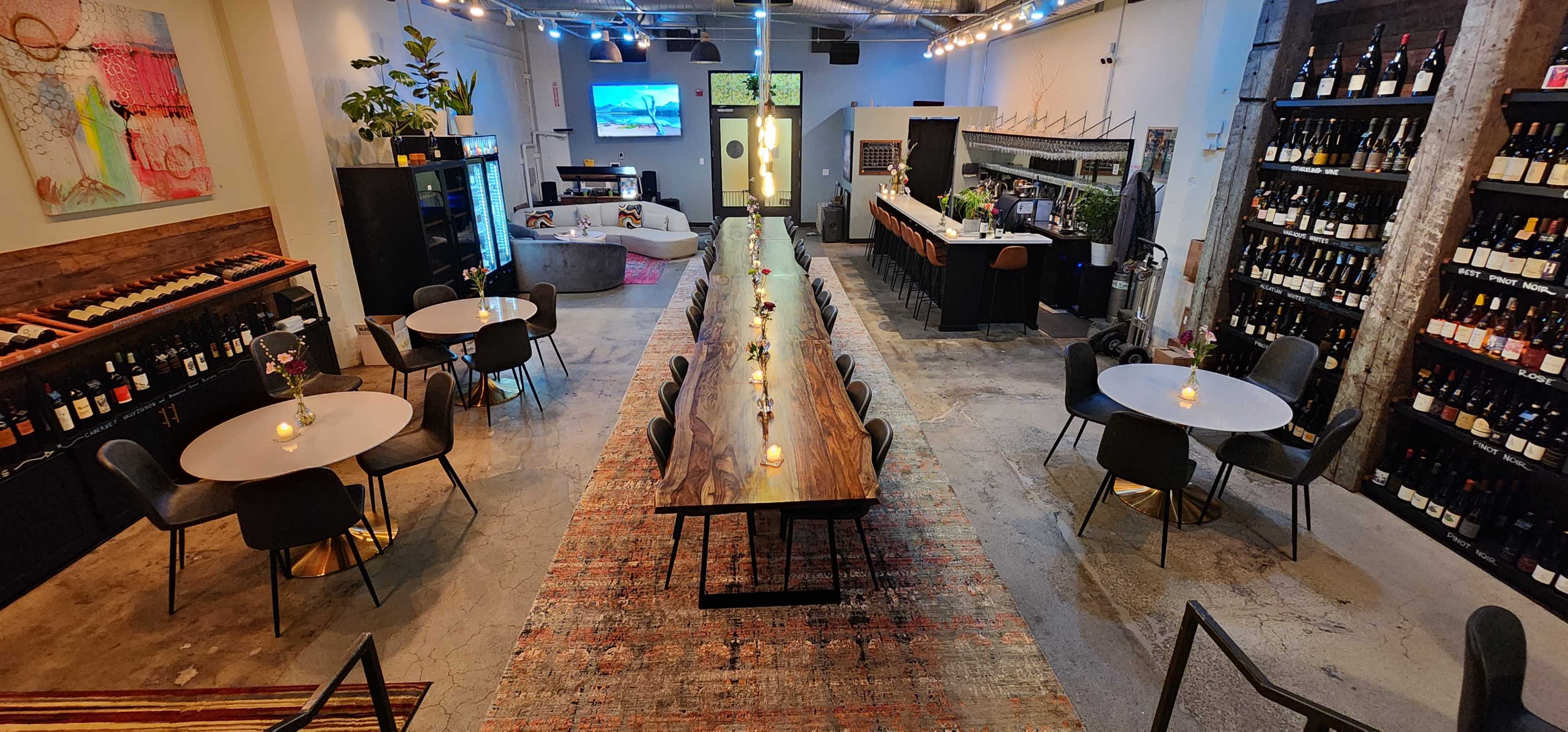 The image shows a spacious interior with a long wooden table surrounded by chairs, several smaller round tables, a bar area, and walls lined with bottles of wine.