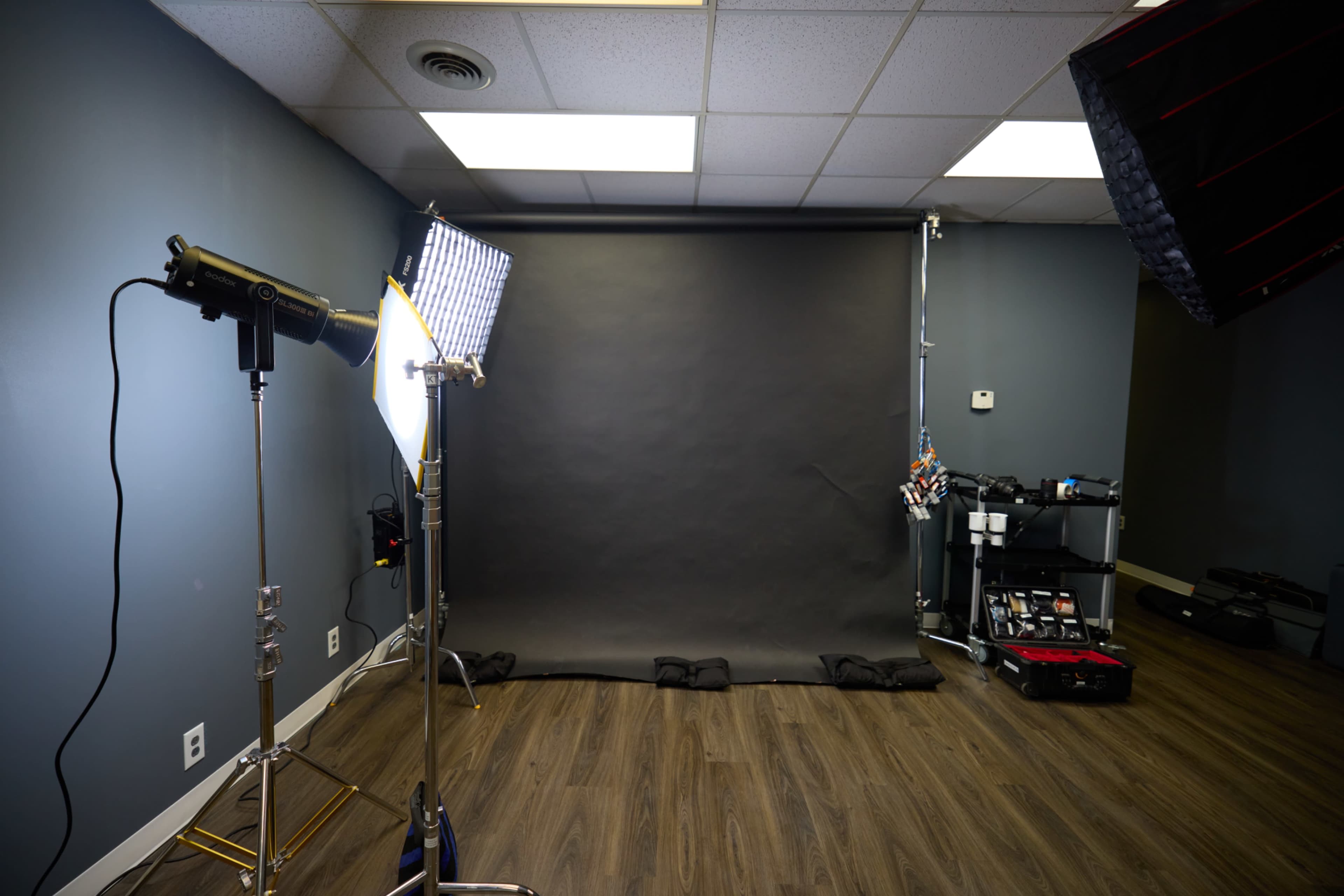 The image shows a photography studio featuring a black backdrop, lighting equipment, and various camera-related accessories.