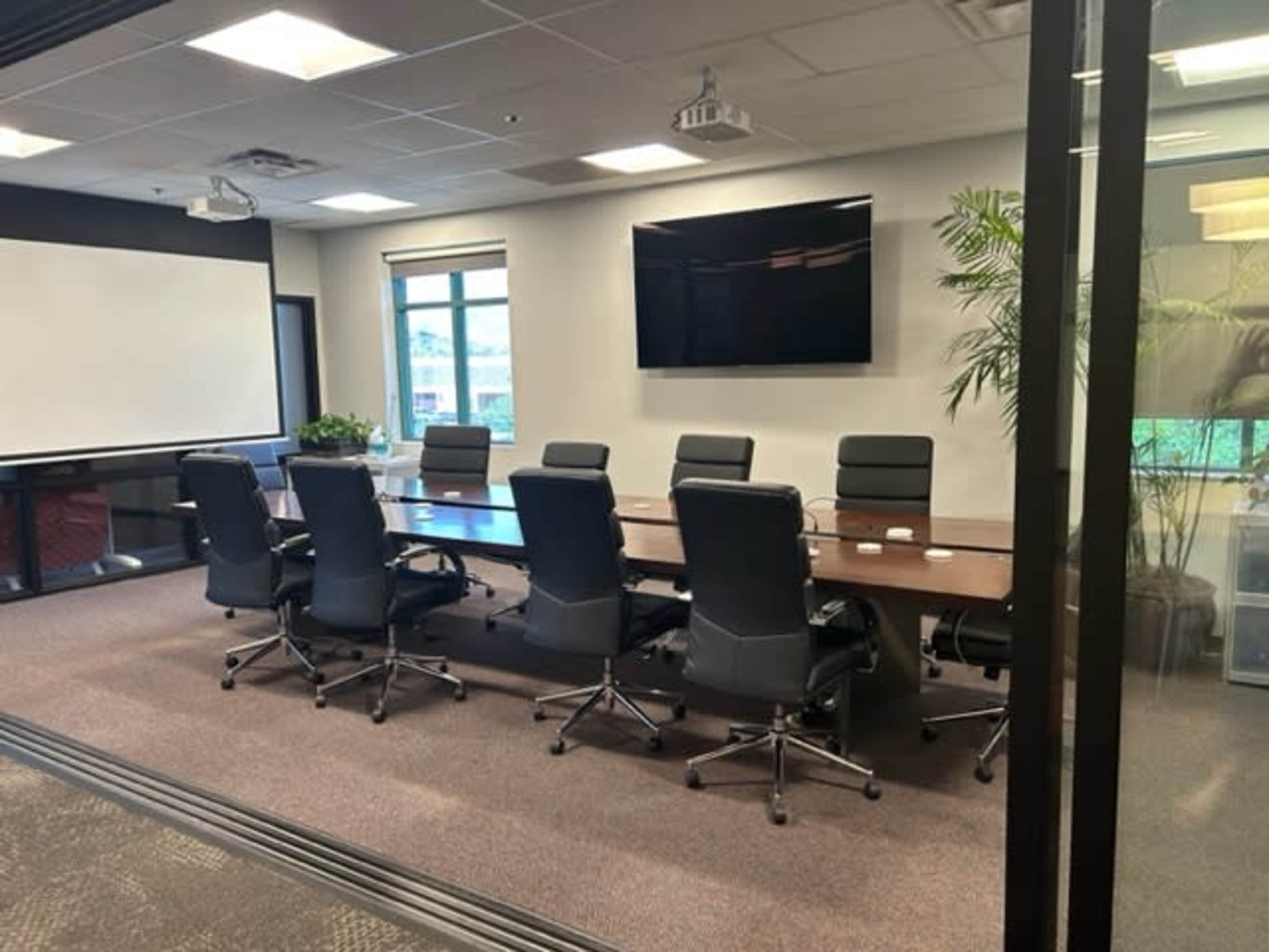 A modern conference room with a long table, eight black chairs, a large screen, and large windows allowing natural light.