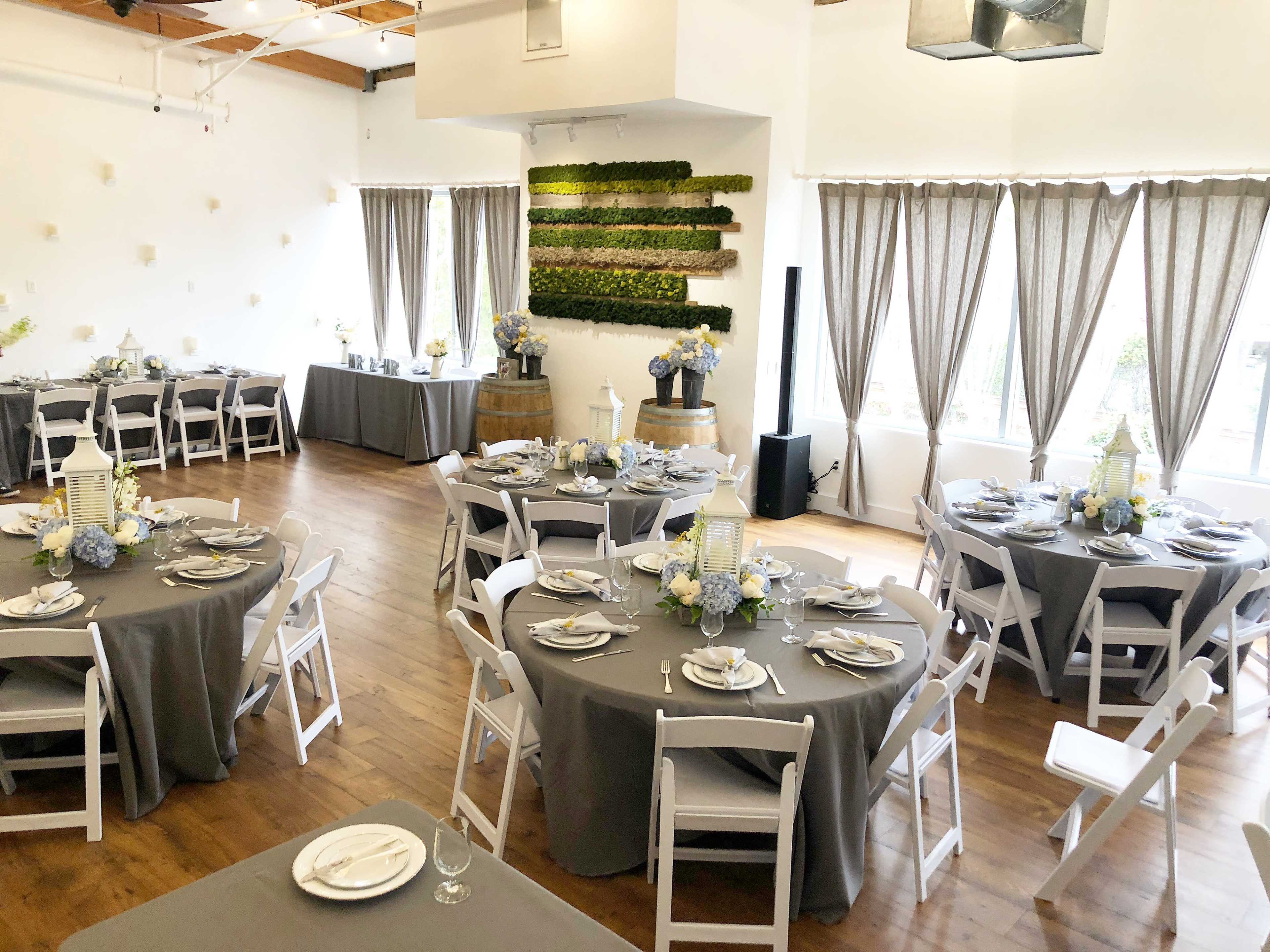 A beautifully arranged event space with round tables covered in gray cloth, each set with white dishes and floral centerpieces, surrounded by wooden flooring and large windows.